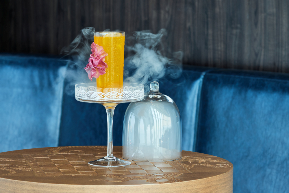 Orange cocktail with pink flower, served on a glass stand, smoke, on wooden table.