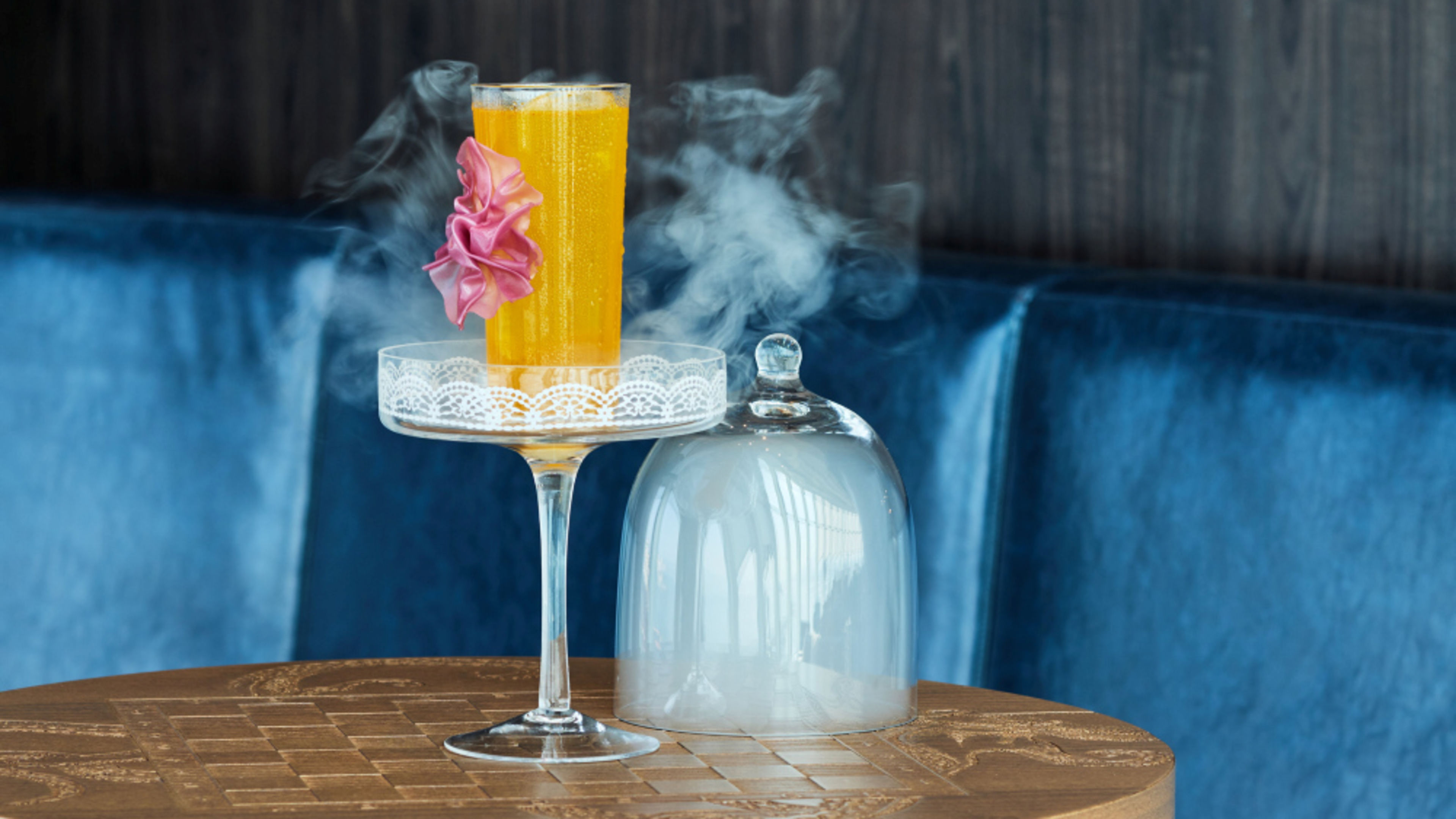 Orange cocktail with pink flower, served on a glass stand, smoke, on wooden table. Orange cocktail with pink flower, served on a glass stand, smoke, on wooden table.