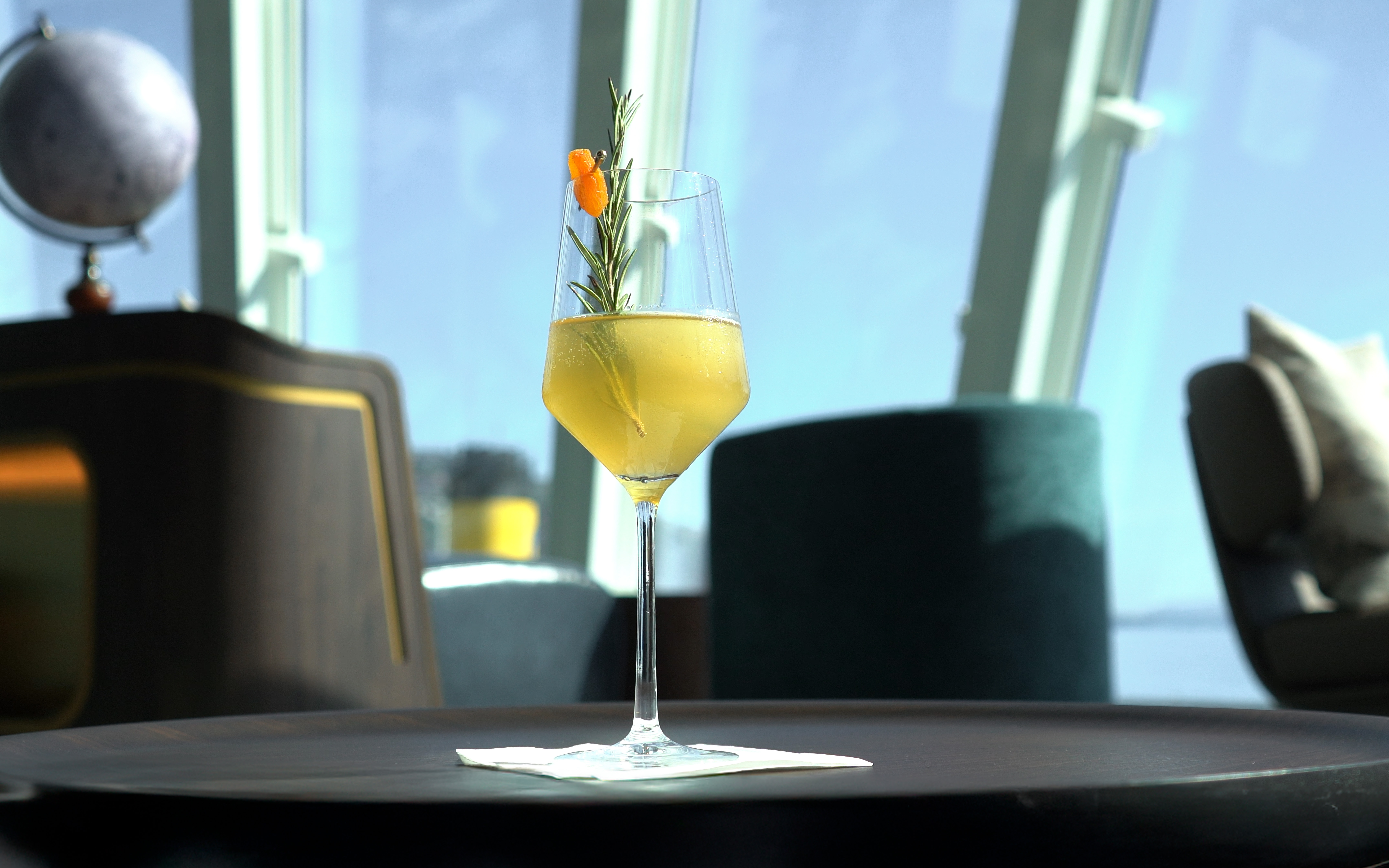 A yellow cocktail in a wine glass, with rosemary and an orange-coloured berry, on a dark table.