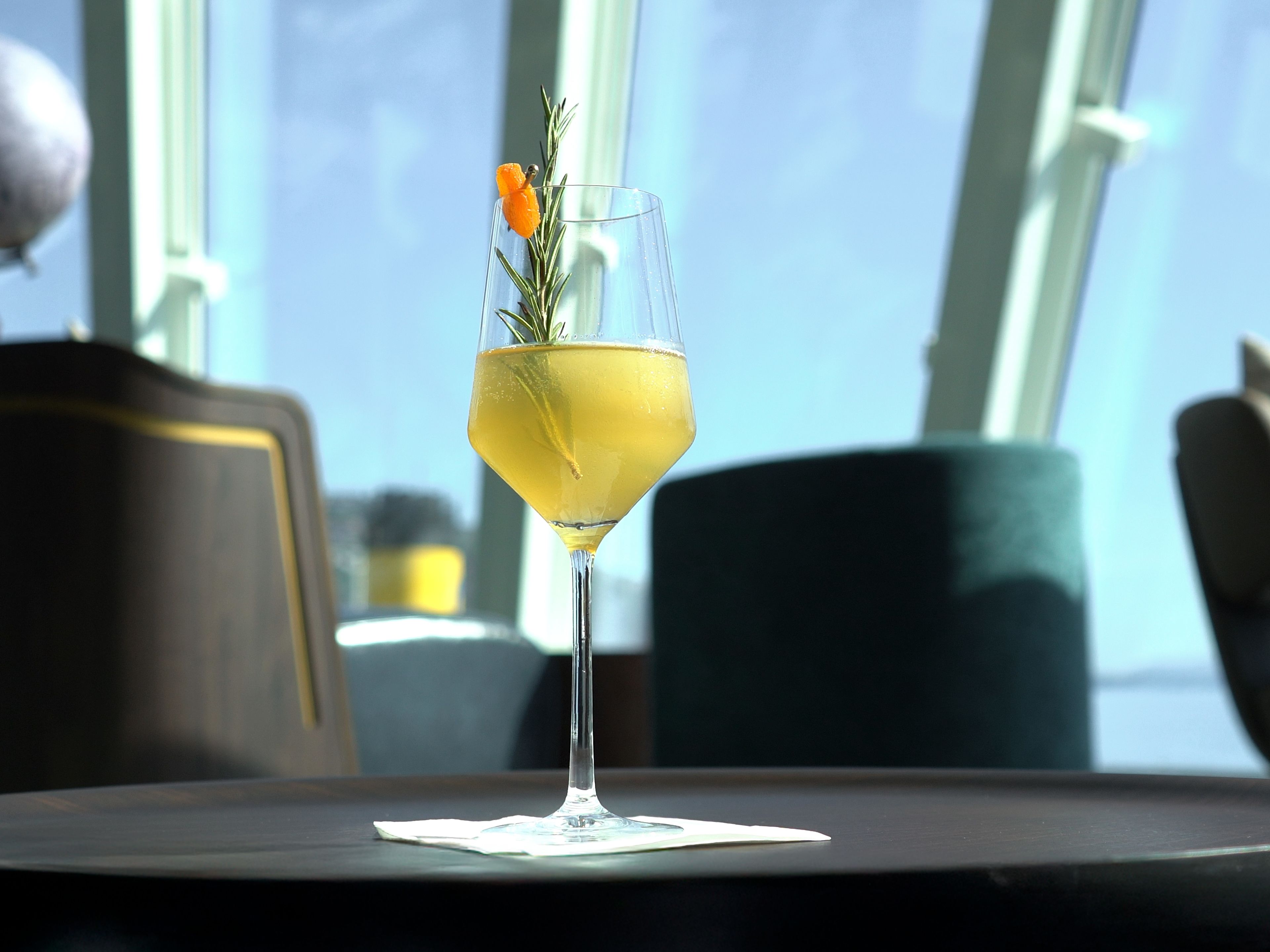A yellow cocktail in a wine glass, with rosemary and an orange-coloured berry, on a dark table. A yellow cocktail in a wine glass, with rosemary and an orange-coloured berry, on a dark table.