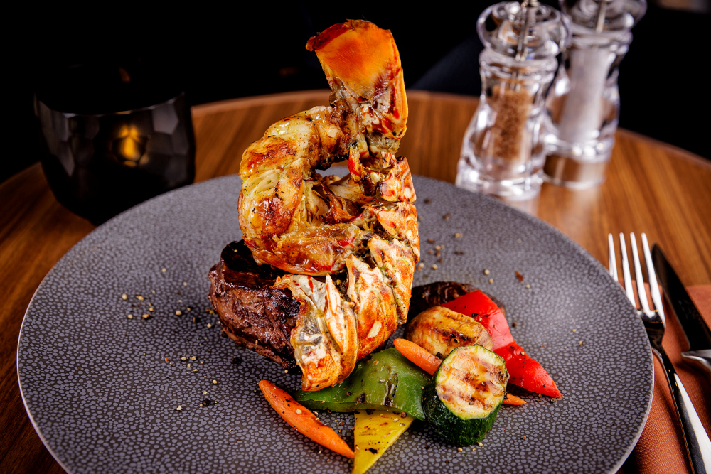 Lobster on steak with grilled vegetables on a grey plate.