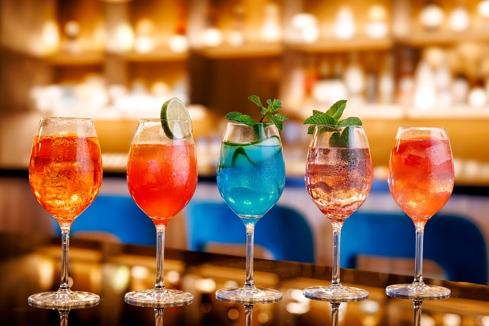 Five colourful Sprizz cocktails in wine glasses, garnished with lime and mint, at a bar.