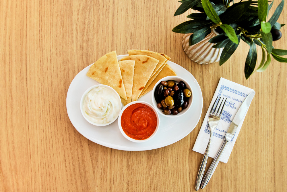 Pita bread with tzatziki, red dip and olives on a wooden table, with cutlery beside it