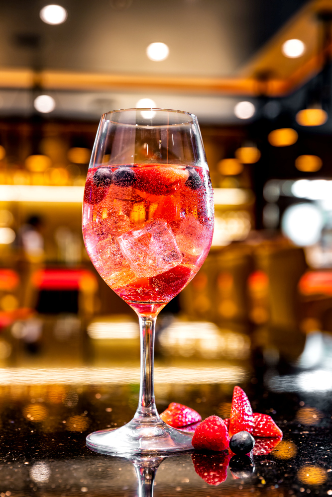A red berry cocktail with ice cubes in a wine glass, with a bar in the background