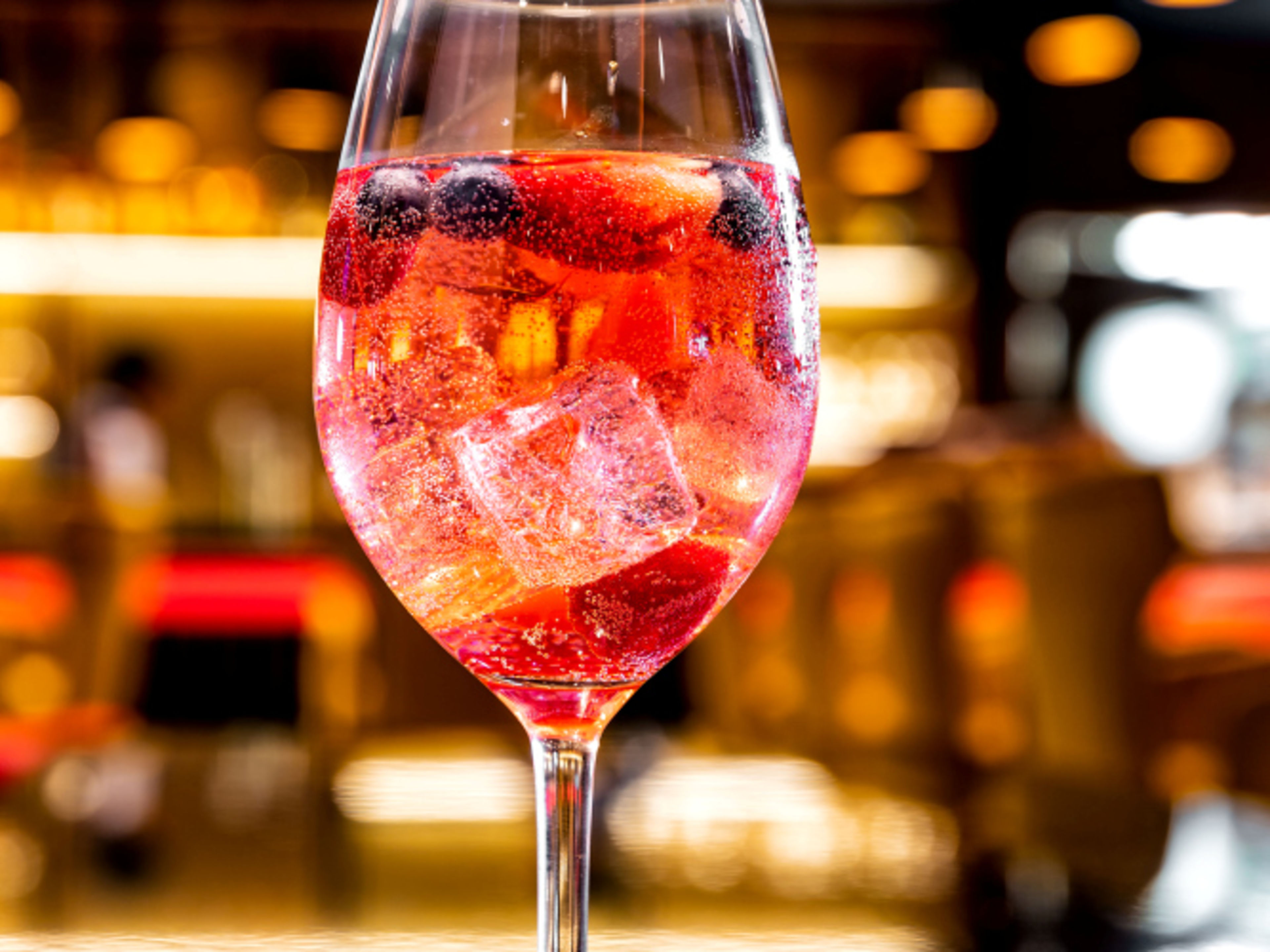 A red berry cocktail with ice cubes in a wine glass, with a bar in the background A red berry cocktail with ice cubes in a wine glass, with a bar in the background