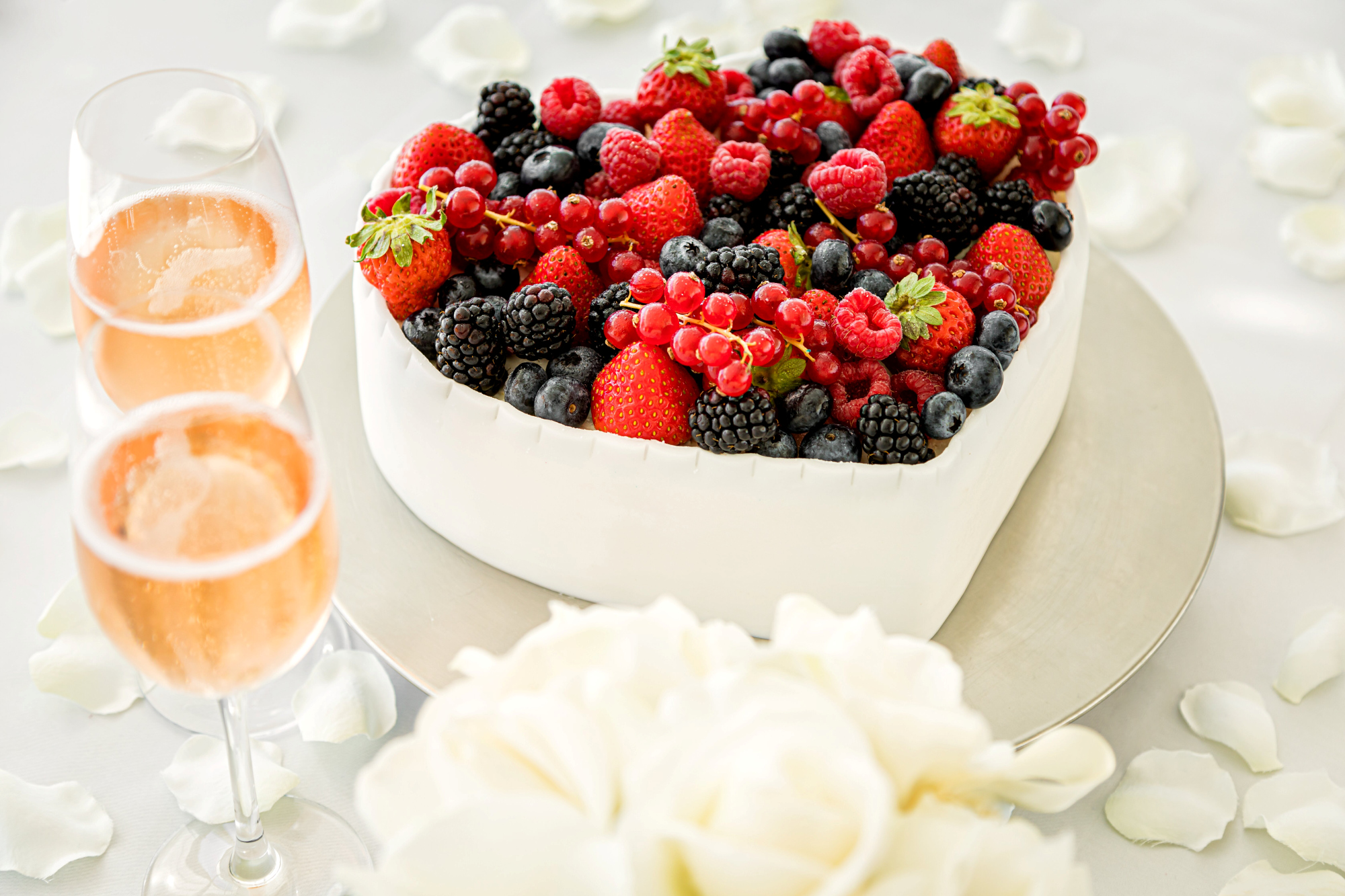 Herzförmige Beerenkuchen mit Sektgläsern und weißen Rosenblättern.