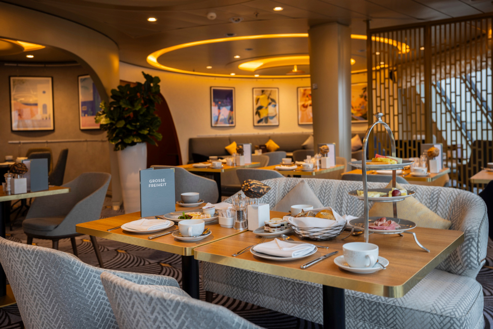 Eleganter Frühstücksbereich in der Café Lounge der Mein Schiff 3, gedeckte Tische, warmes Licht, graue Sitzbänke und Kunstbilder an der Wand