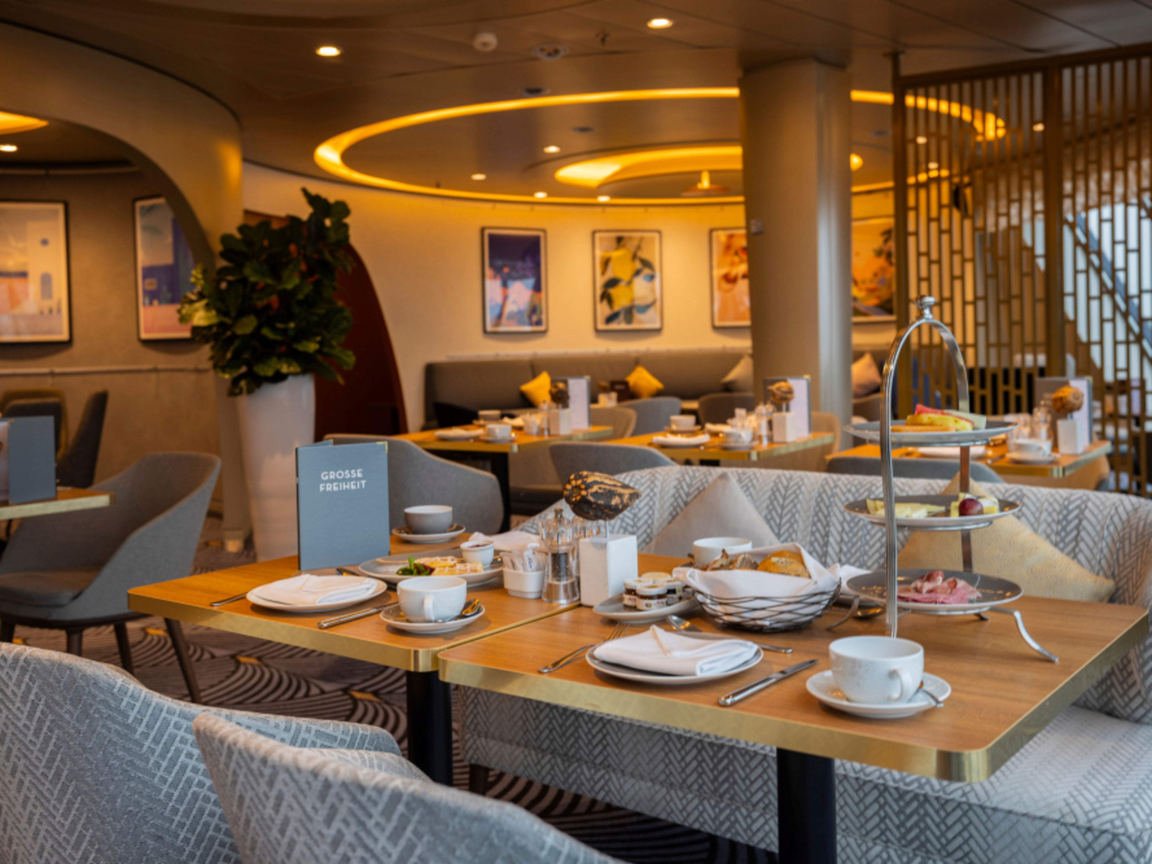 Eleganter Frühstücksbereich in der Café Lounge der Mein Schiff 3, gedeckte Tische, warmes Licht, graue Sitzbänke und Kunstbilder an der Wand Eleganter Frühstücksbereich in der Café Lounge der Mein Schiff 3, gedeckte Tische, warmes Licht, graue Sitzbänke und Kunstbilder an der Wand