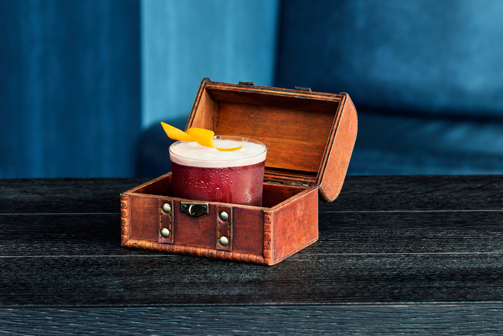 A dark red drink with foam and orange zest, served in a glass inside an open treasure chest.