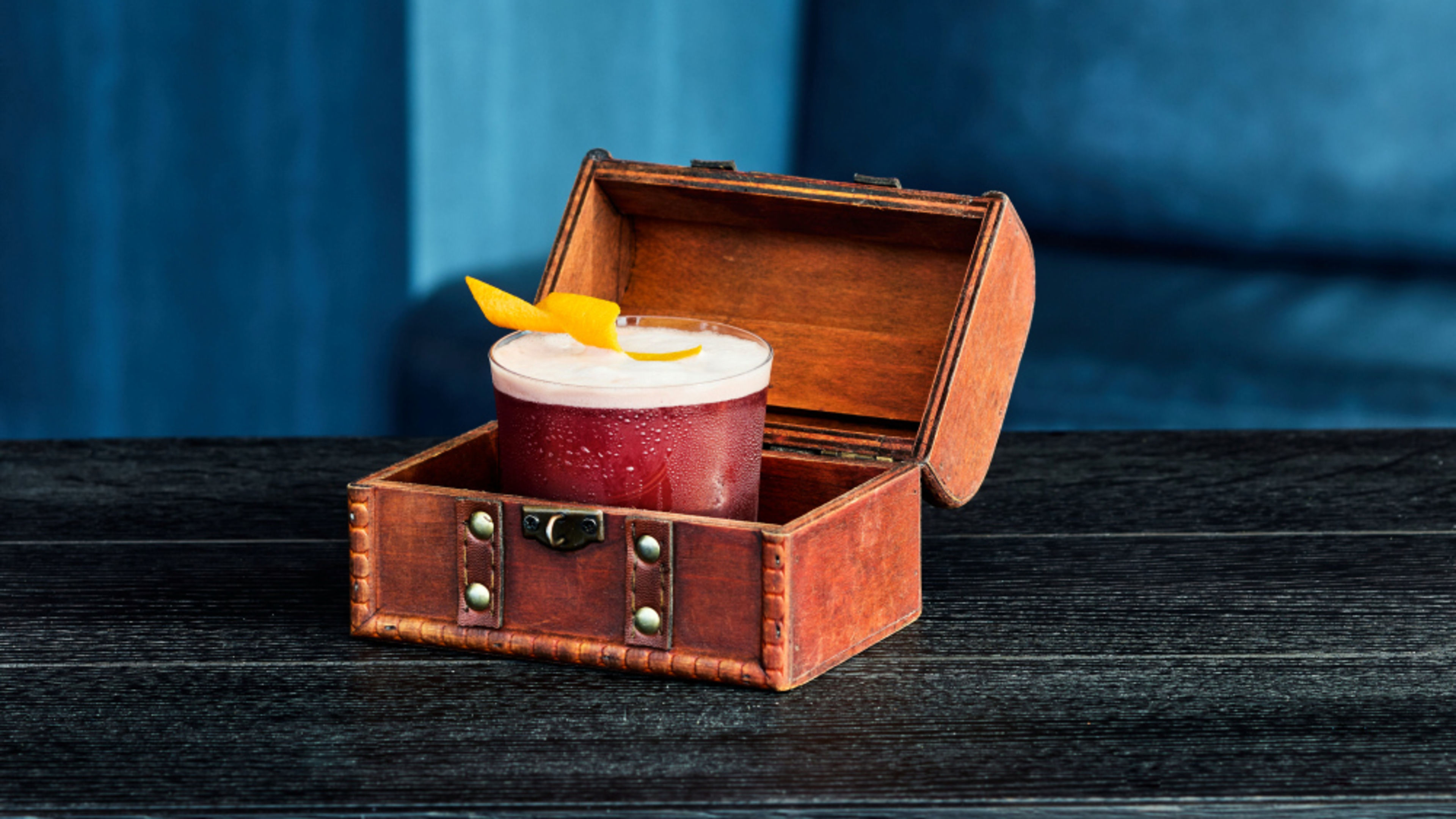 A dark red drink with foam and orange zest, served in a glass inside an open treasure chest. A dark red drink with foam and orange zest, served in a glass inside an open treasure chest.