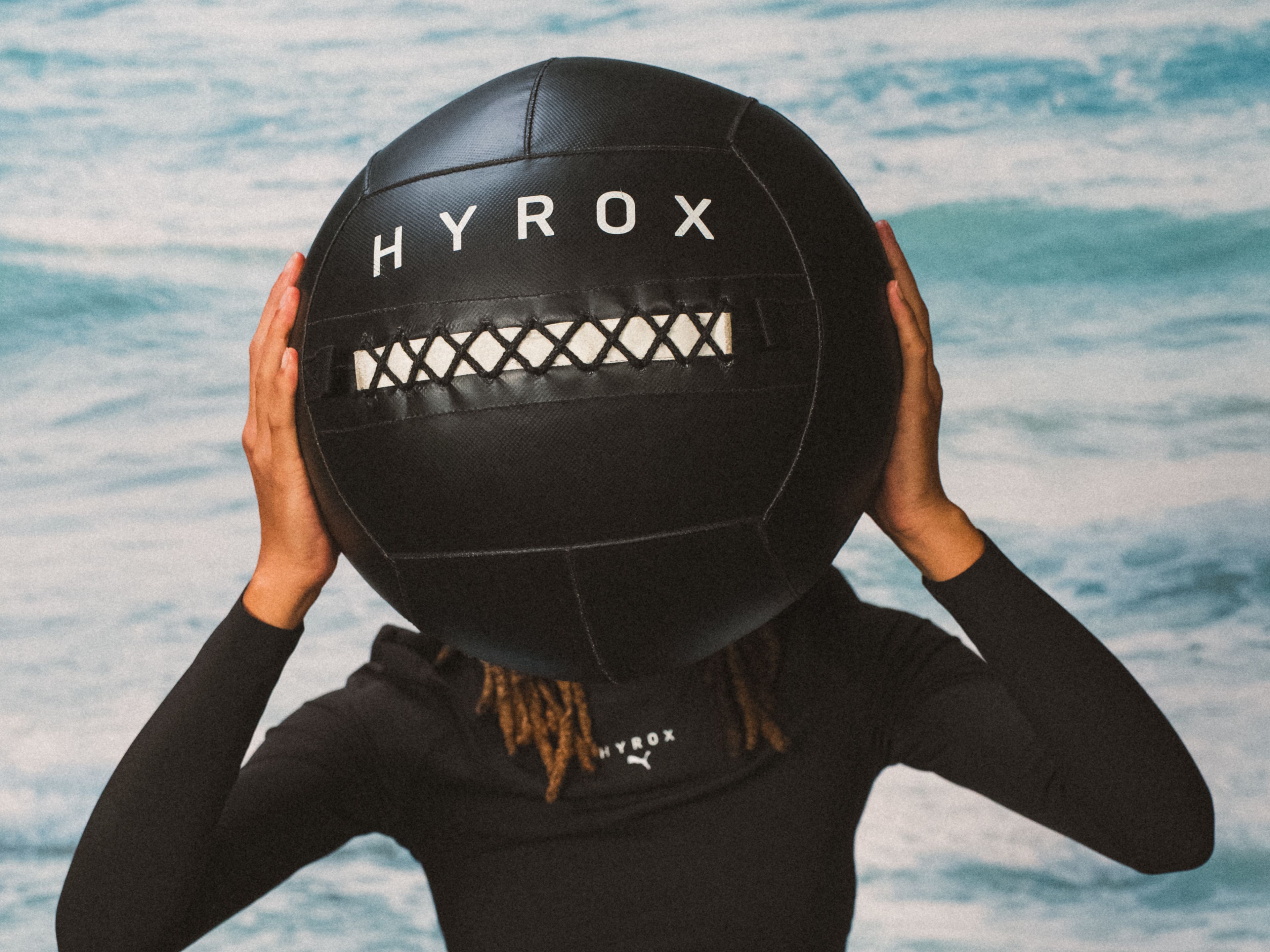 Person holding large black Hyrox ball in front of blue background. Person holding large black Hyrox ball in front of blue background.