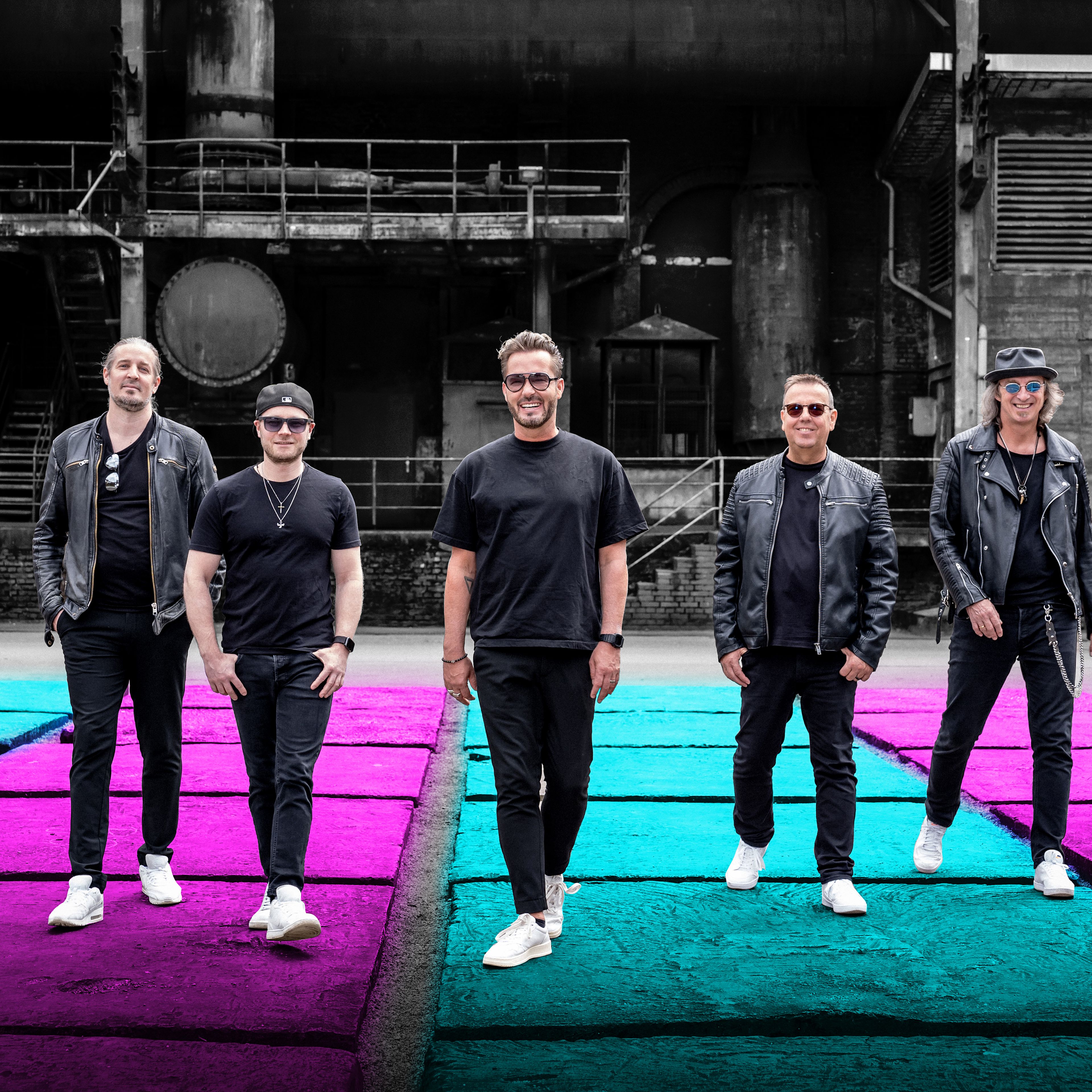 Fünf Männer in schwarzen Outfits stehen auf buntem Boden vor Industriekulisse. Fünf Männer in schwarzen Outfits stehen auf buntem Boden vor Industriekulisse.