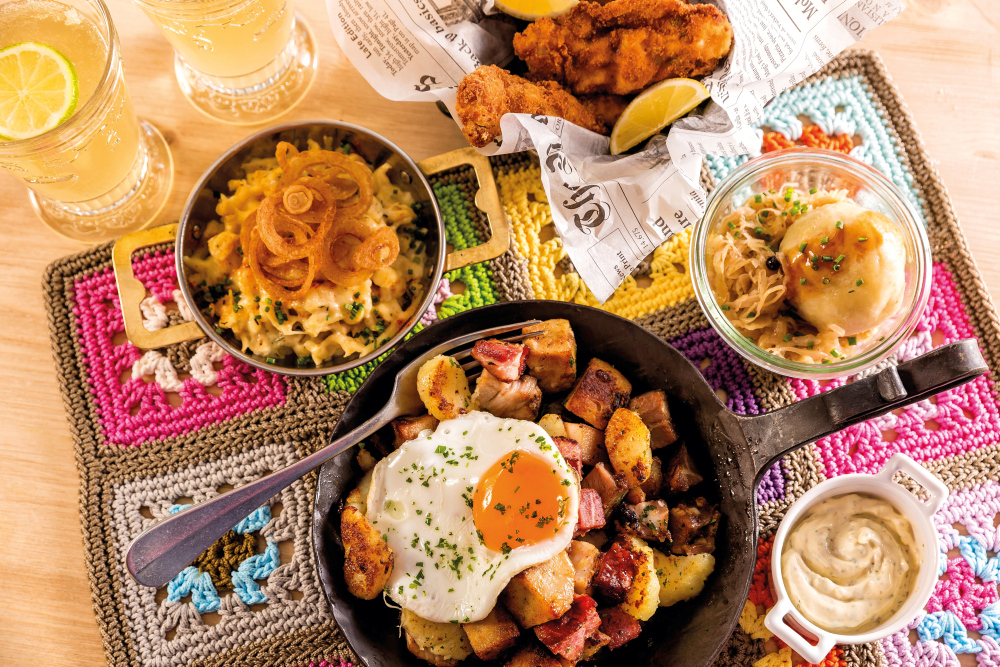 A hearty brunch with fried eggs, fried potatoes, cheese spaetzle, sauerkraut and drinks, served on a colourful placemat.