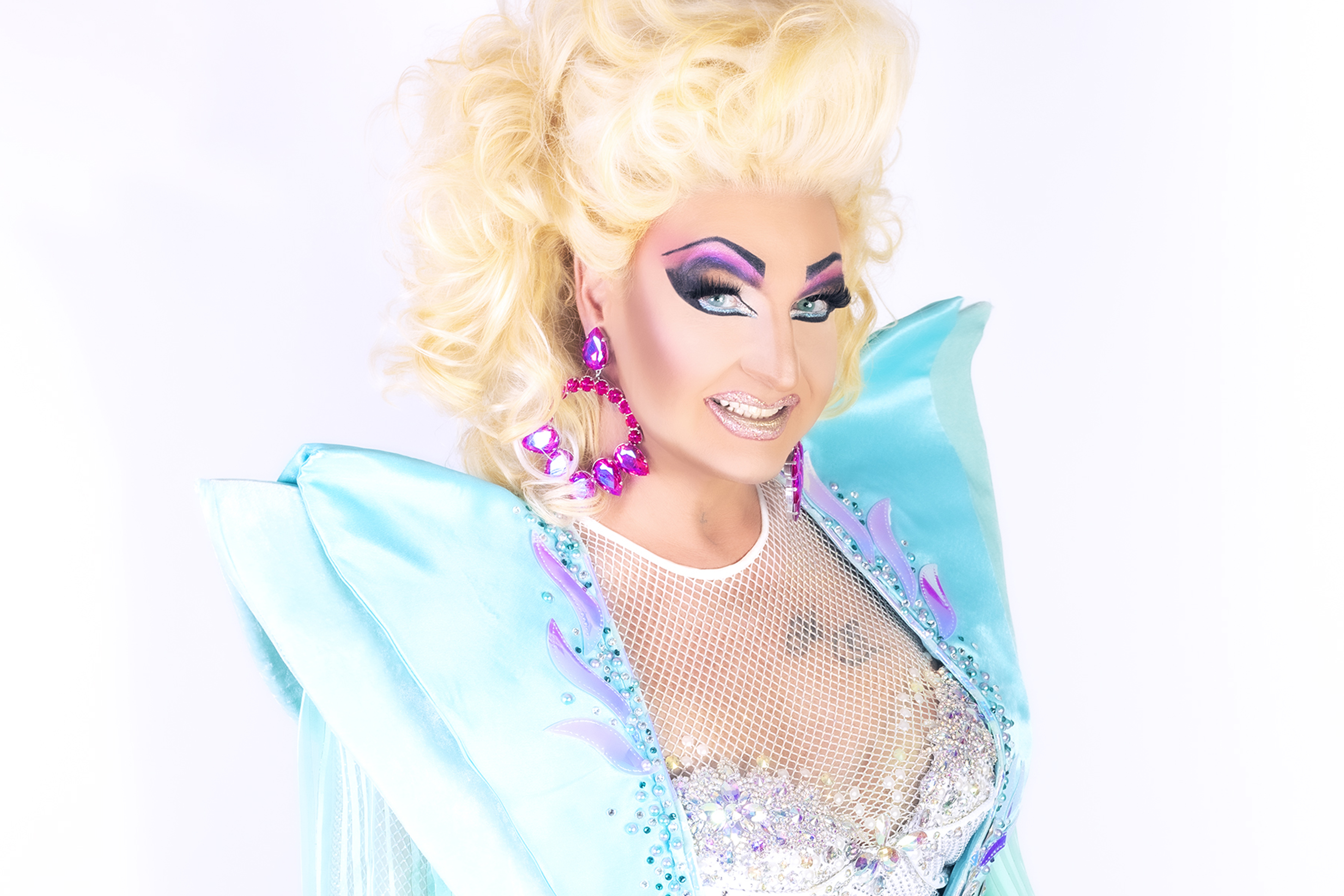 Drag Queen in türkisem Kostüm, blonder Perücke und pinken Ohrringen lächelt glamourös.