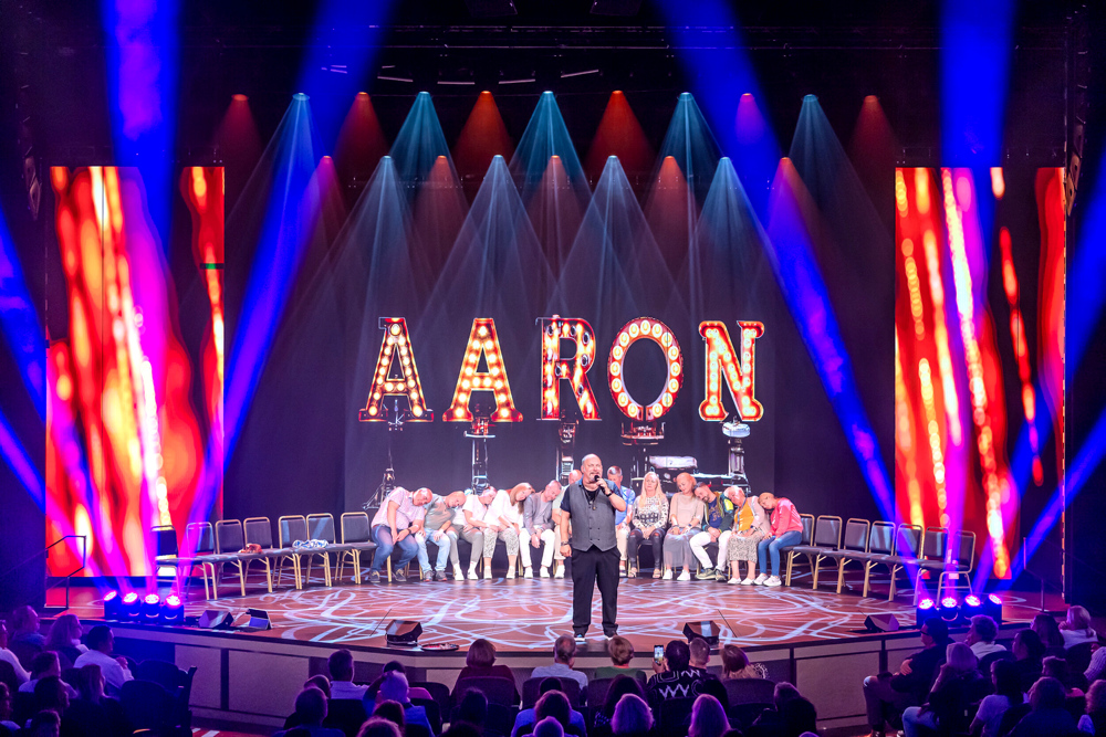Mann auf Bühne mit leuchtendem "AARON"-Schild, Publikum schaut zu.