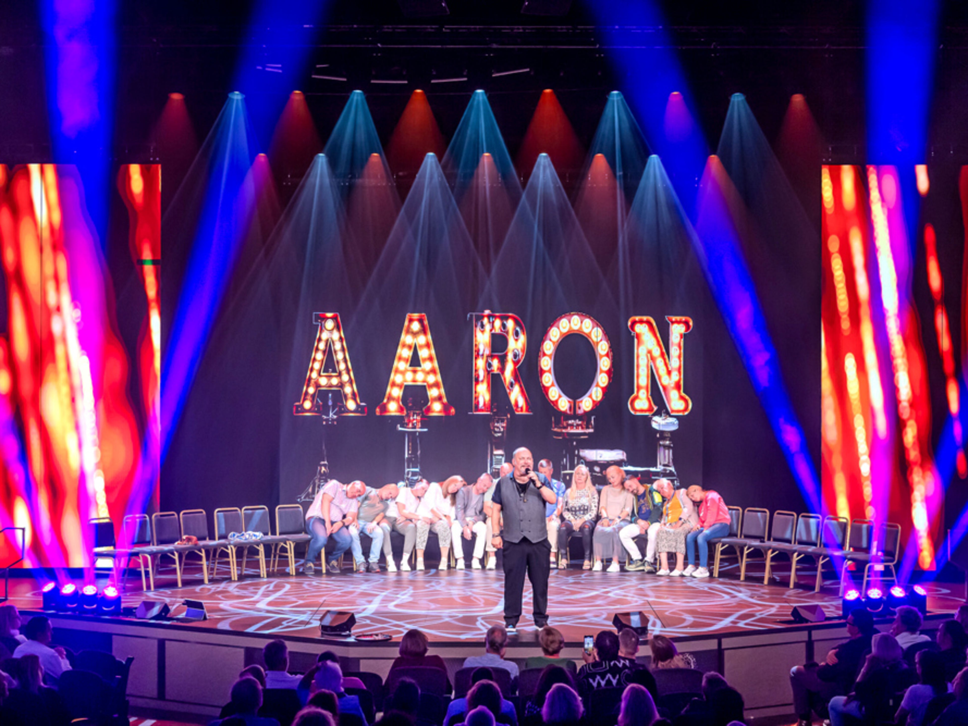 Mann auf Bühne mit leuchtendem "AARON"-Schild, Publikum schaut zu. Mann auf Bühne mit leuchtendem "AARON"-Schild, Publikum schaut zu.