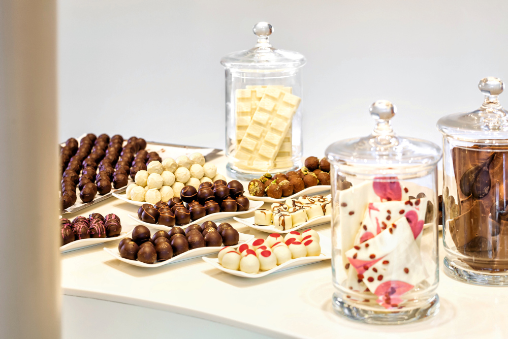 Elegante Pralinenteller und Glasgefäße mit weißer und dunkler Schokolade auf weißem Tisch.