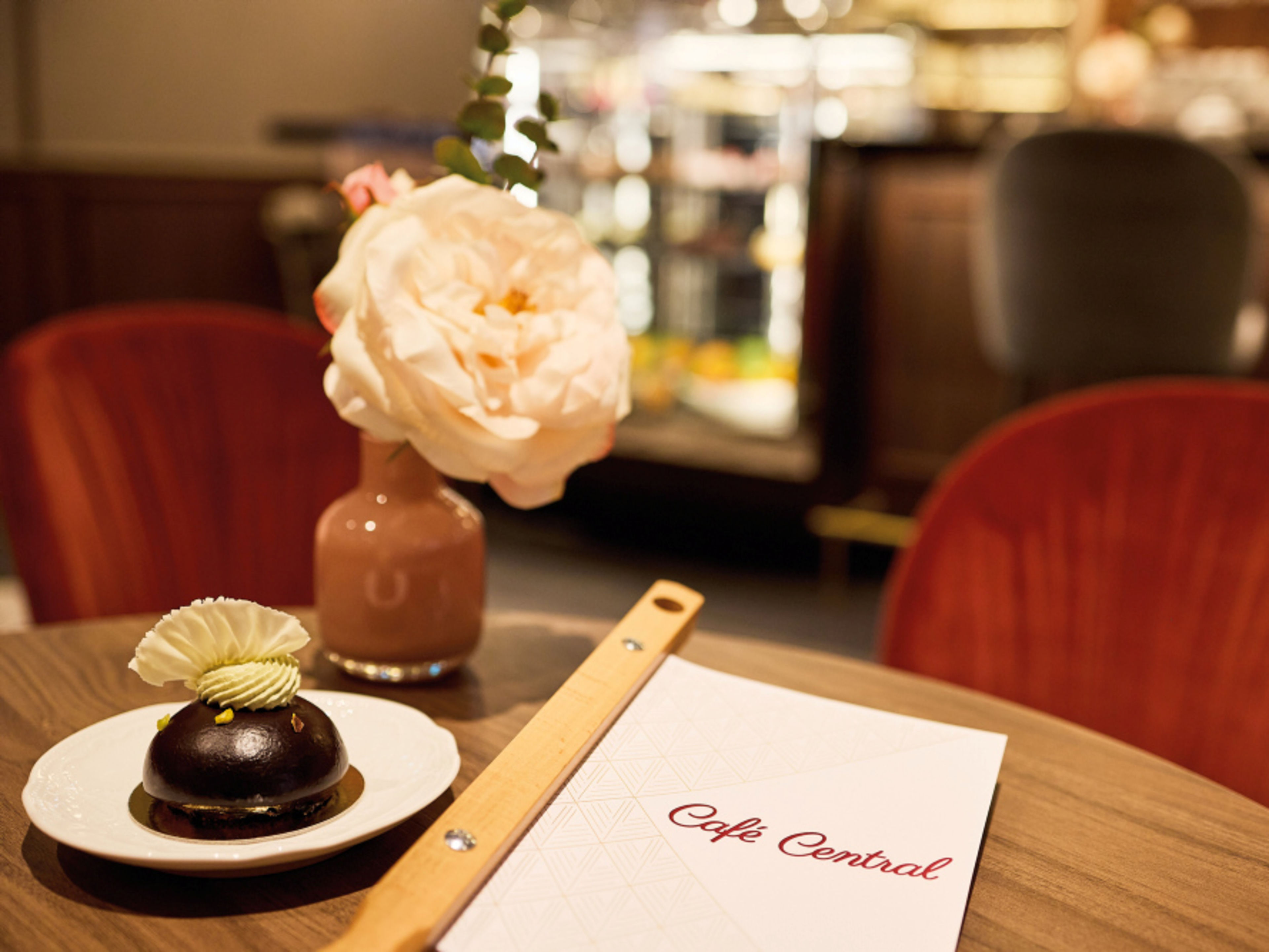 Elegantes Café mit Schokoladendessert, Speisekarte und rosa Rosenblüte in Vase auf Holztisch mit roten Samtstühlen Elegantes Café mit Schokoladendessert, Speisekarte und rosa Rosenblüte in Vase auf Holztisch mit roten Samtstühlen