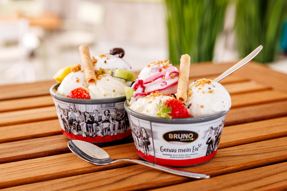 Zwei Becher Eiscreme mit Früchten und Waffeln auf Holztisch.