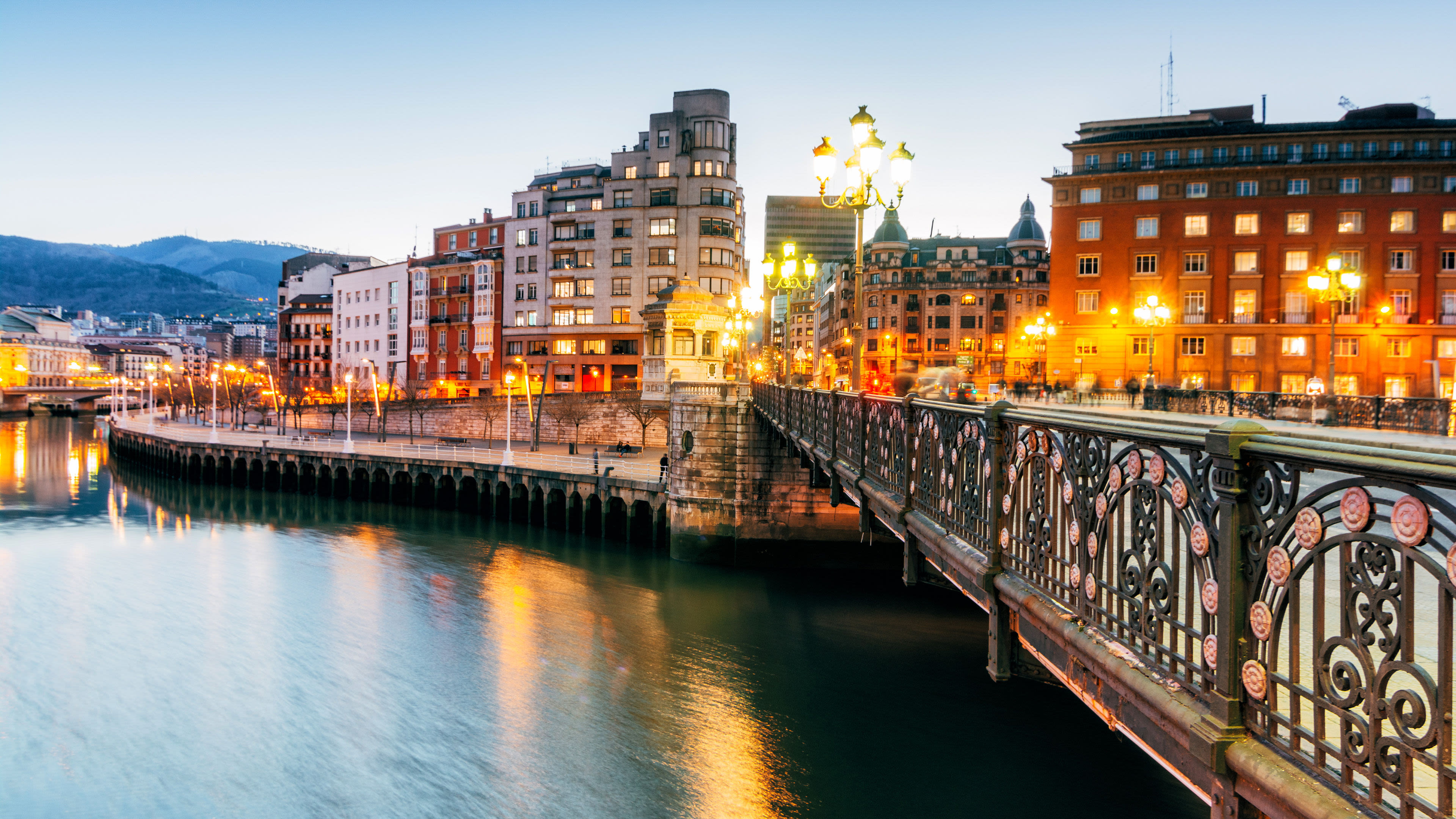Brücke und beleuchtete Gebäude am Flussufer in Bilbao bei Dämmerung.