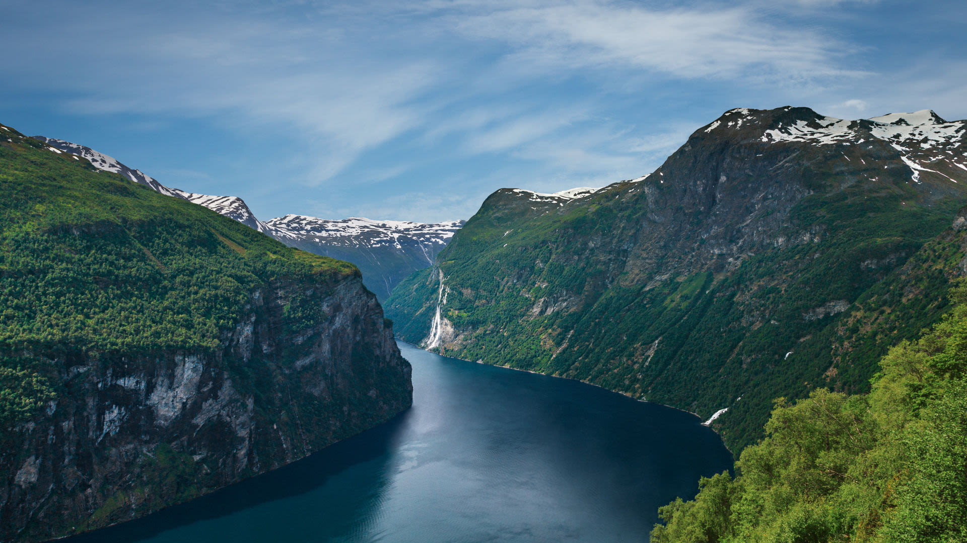 Norwegischer Fjord mit Wasserfall, umgeben von grünen Bergen und blauem Himmel.