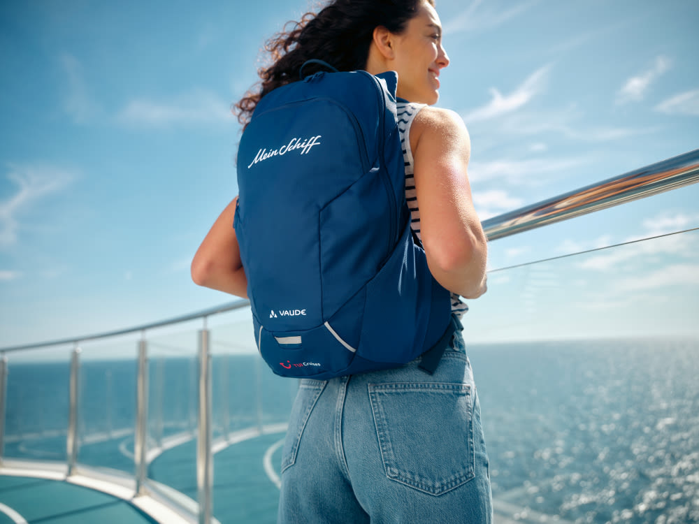 Frau mit blauem Rucksack auf Kreuzfahrtschiff, Meer im Hintergrund.