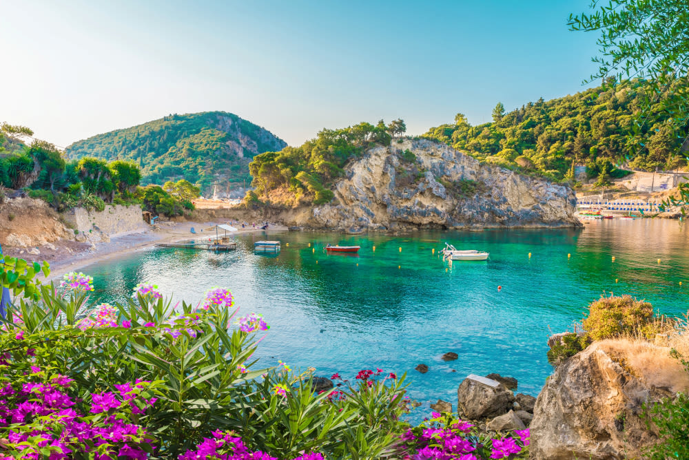 Bucht auf Korfu mit türkisfarbenem Wasser, Booten und Blumen.