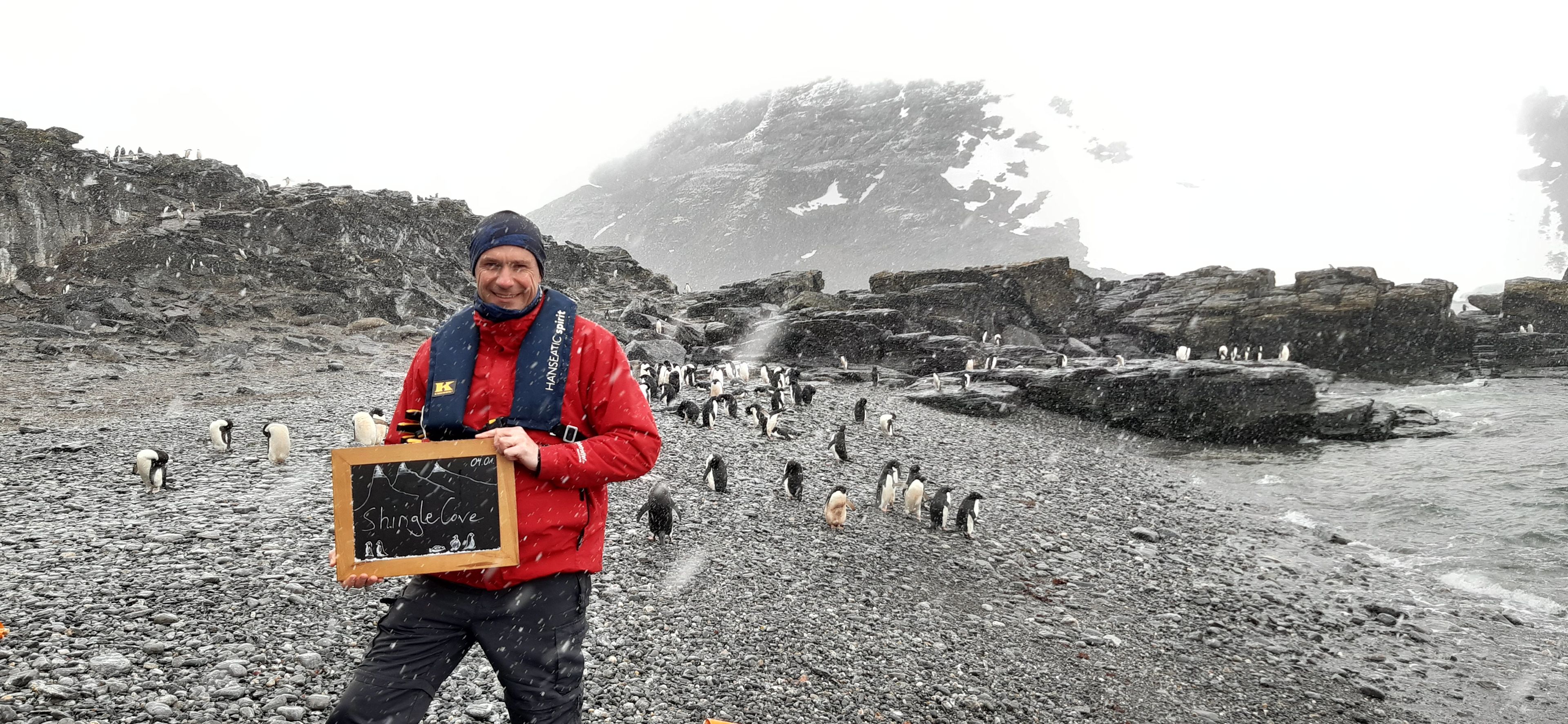 Mann in roter Jacke mit Pinguinen am verschneiten Strand von Shingle Cove.