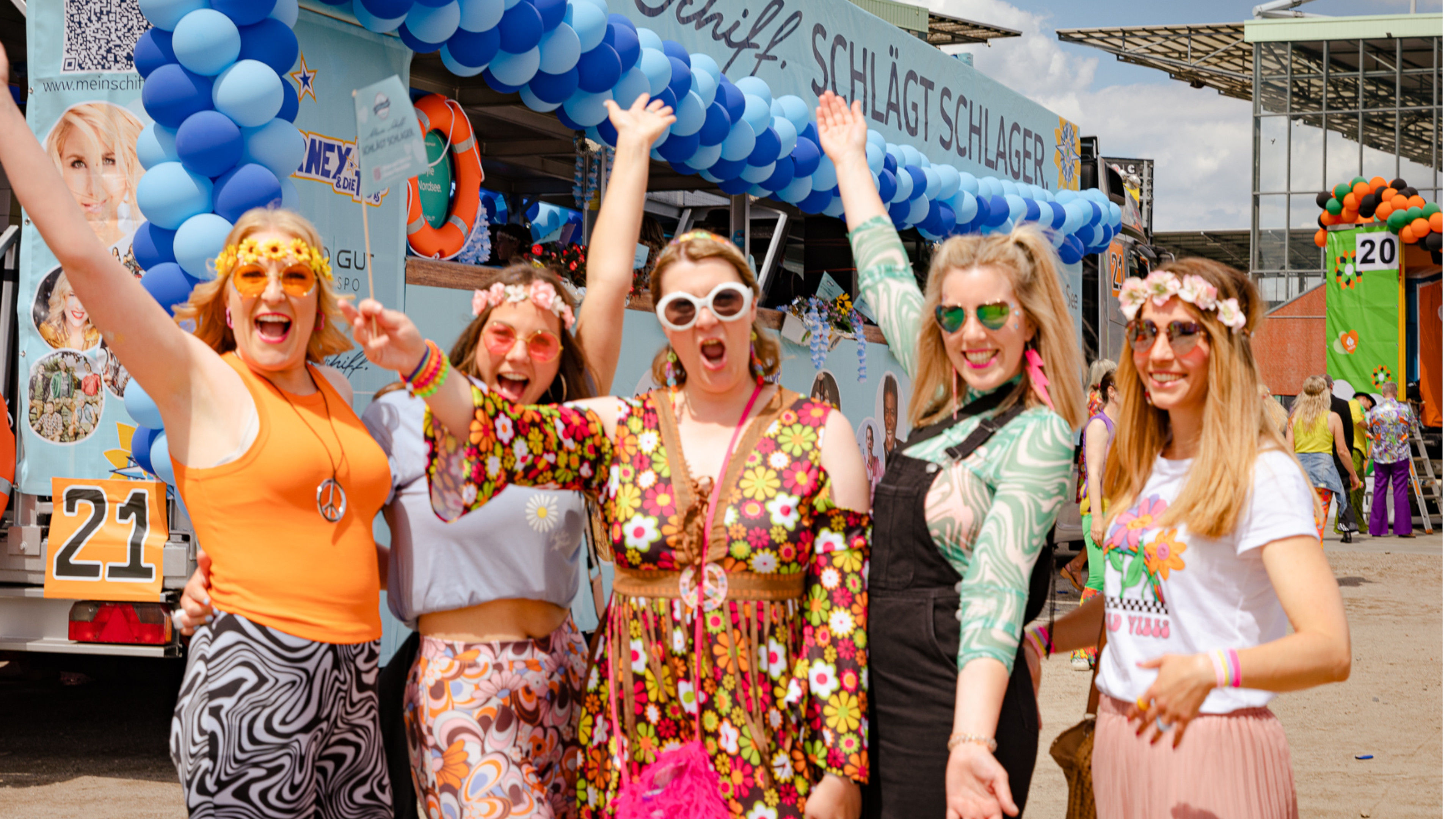 Gruppe fröhlicher Frauen in bunter Hippie-Kleidung vor geschmücktem Wagen.