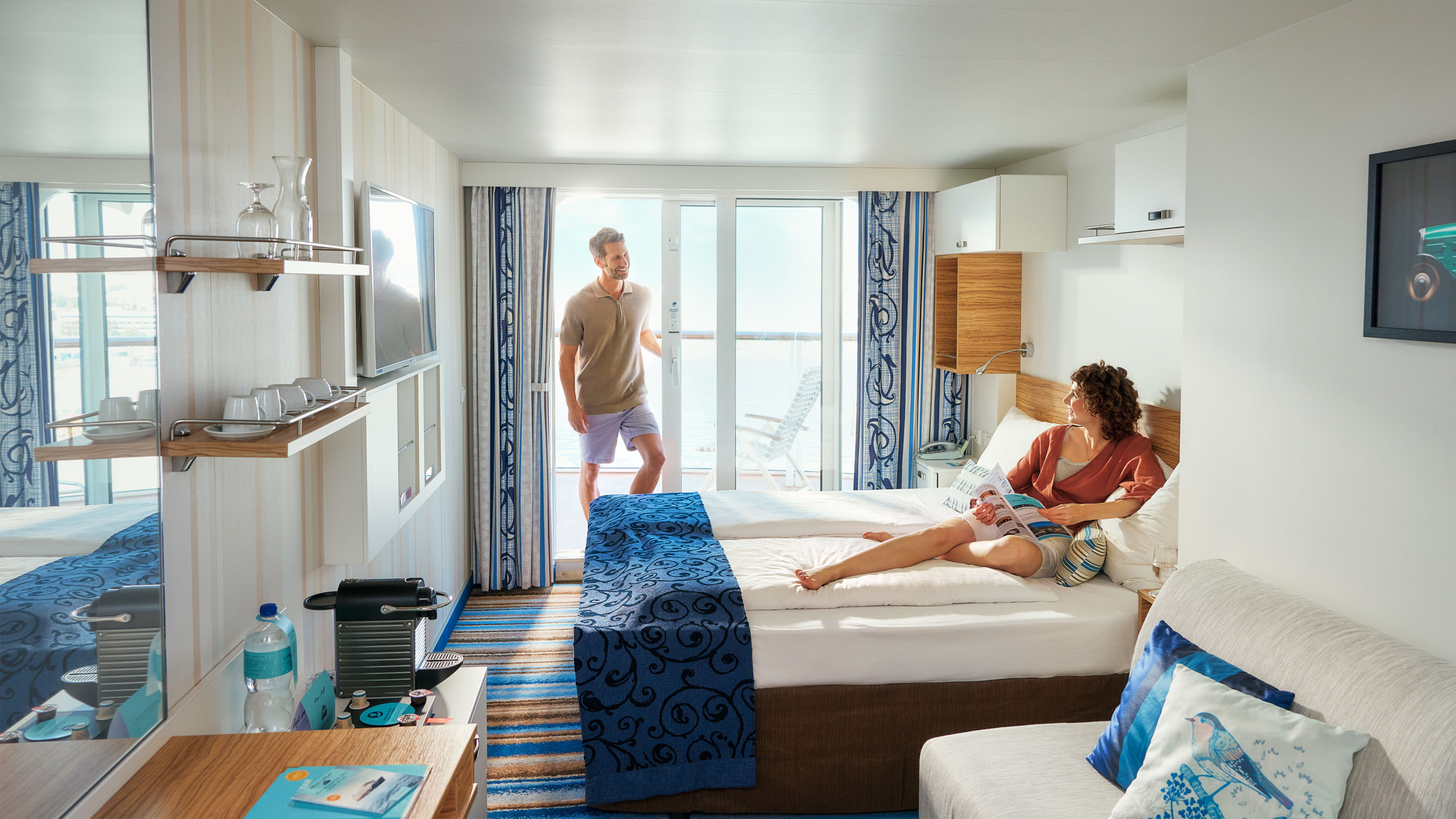 Paar entspannt in heller Kreuzfahrtkabine mit Balkon.