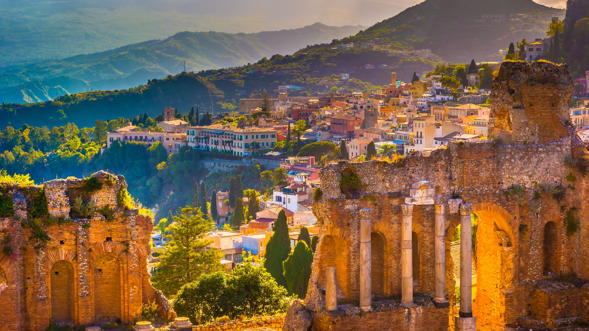 Antike Ruinen in Taormina, Sizilien, mit bunten Häusern und grünen Hügeln im Hintergrund.
