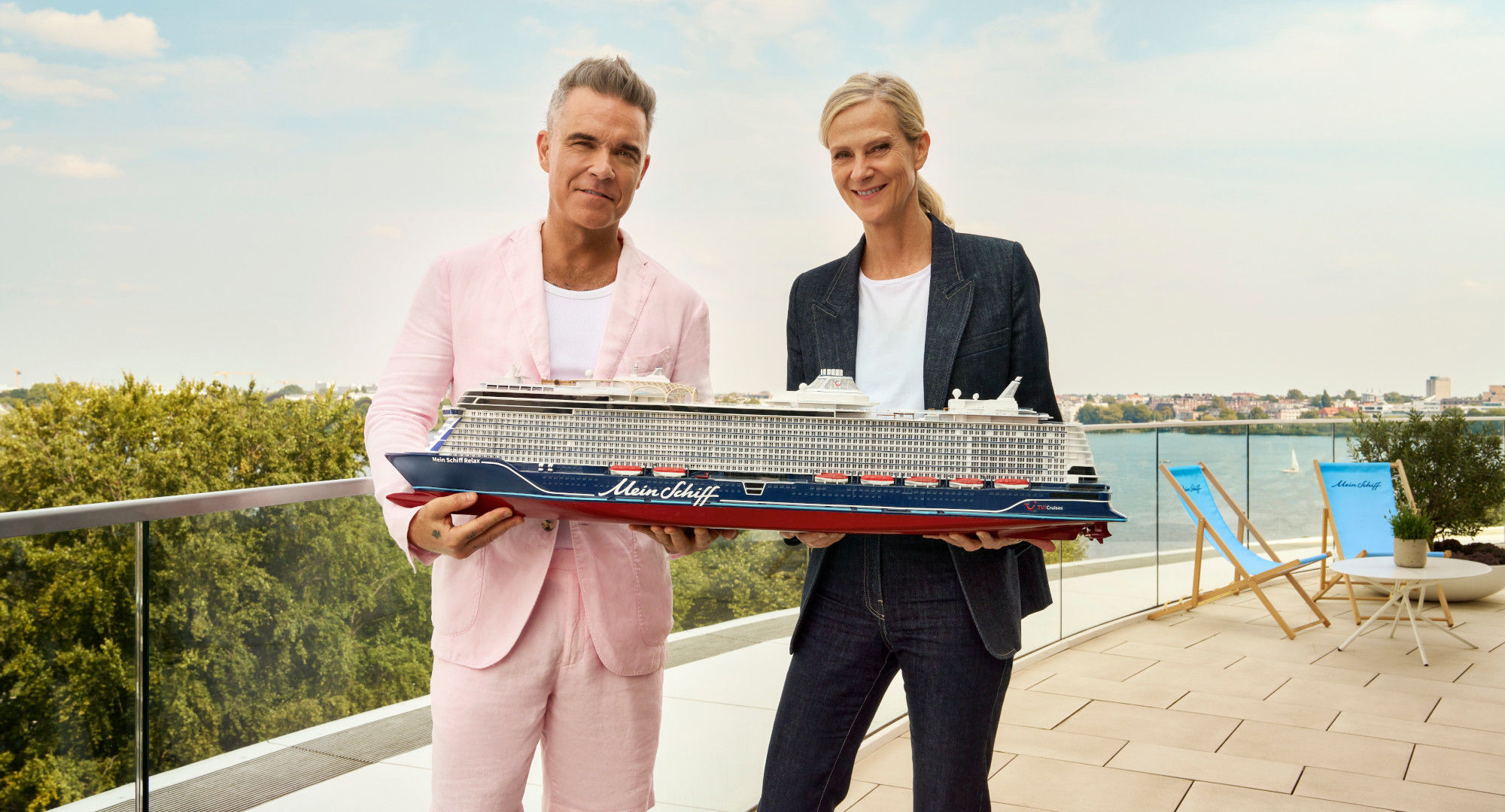 Zwei Personen halten Modell eines Kreuzfahrtschiffs der Mein Schiff® Flotte auf Terrasse mit Seeblick.