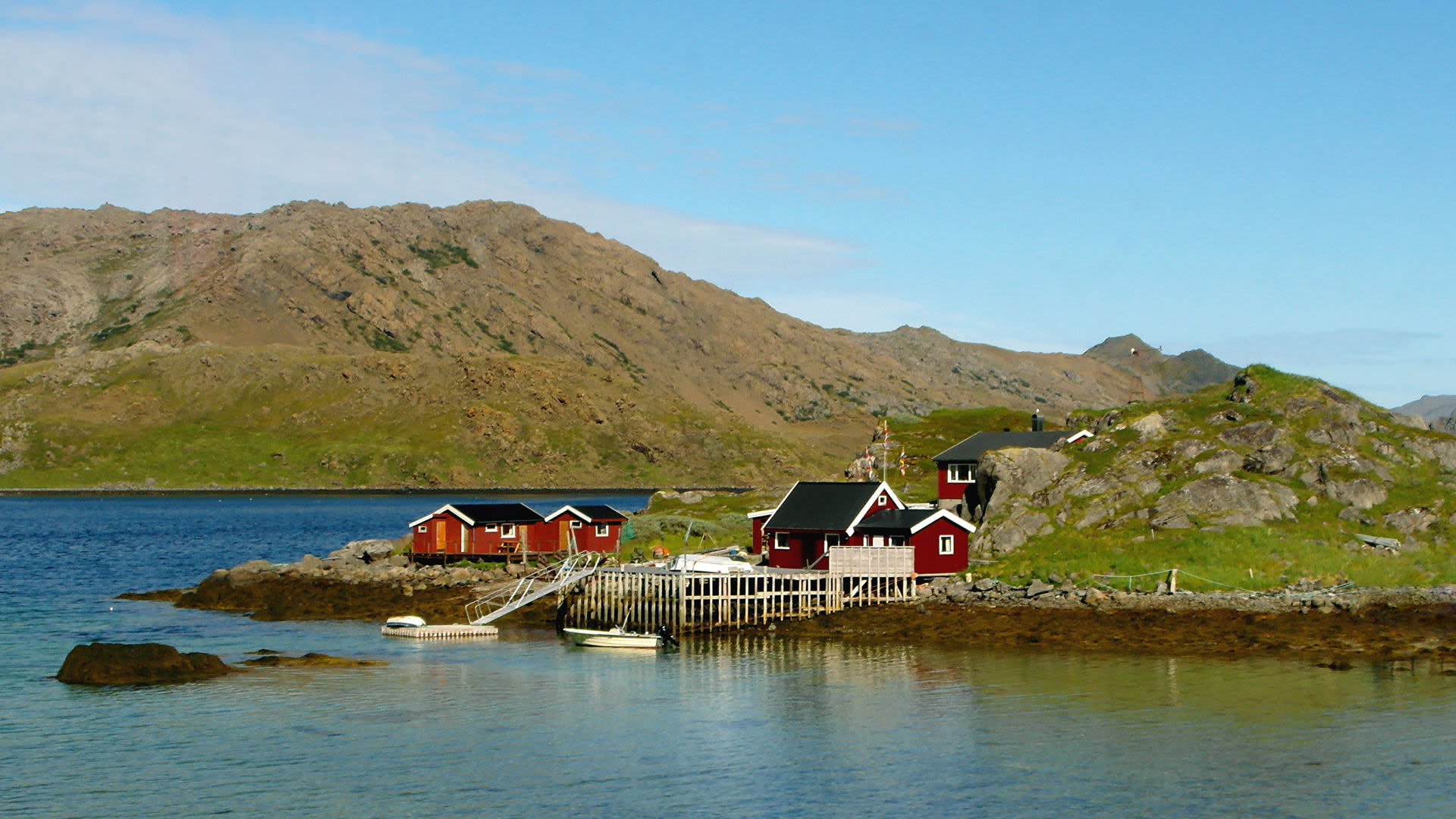 Rote Hütten am Fjord, umgeben von Bergen und blauem Himmel.