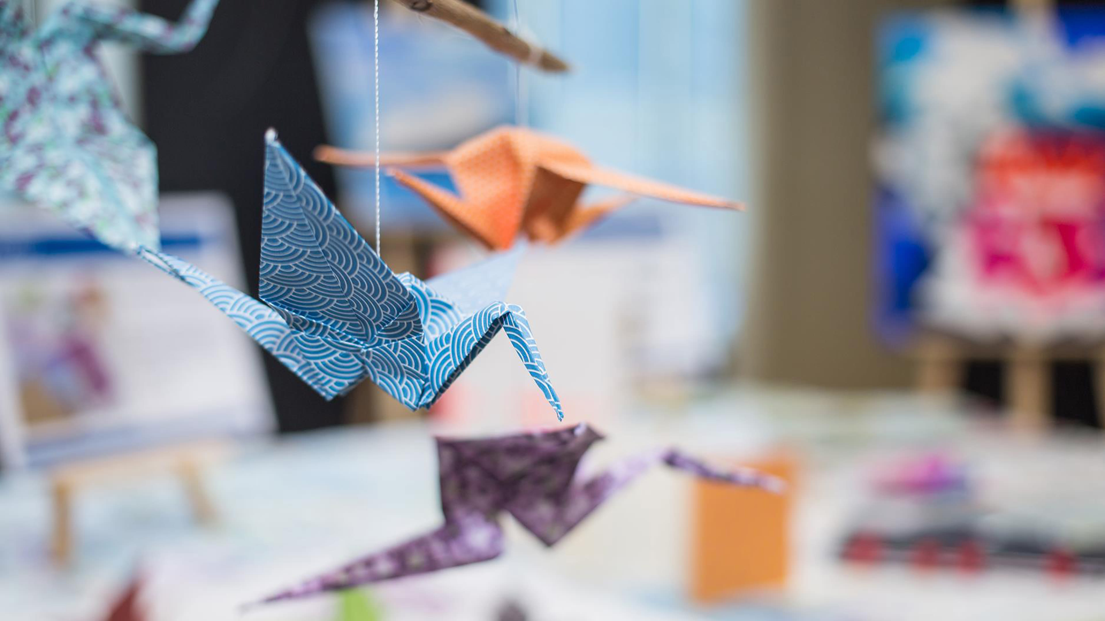 Bunte Origami-Kraniche hängen an Fäden, unscharfer Hintergrund.