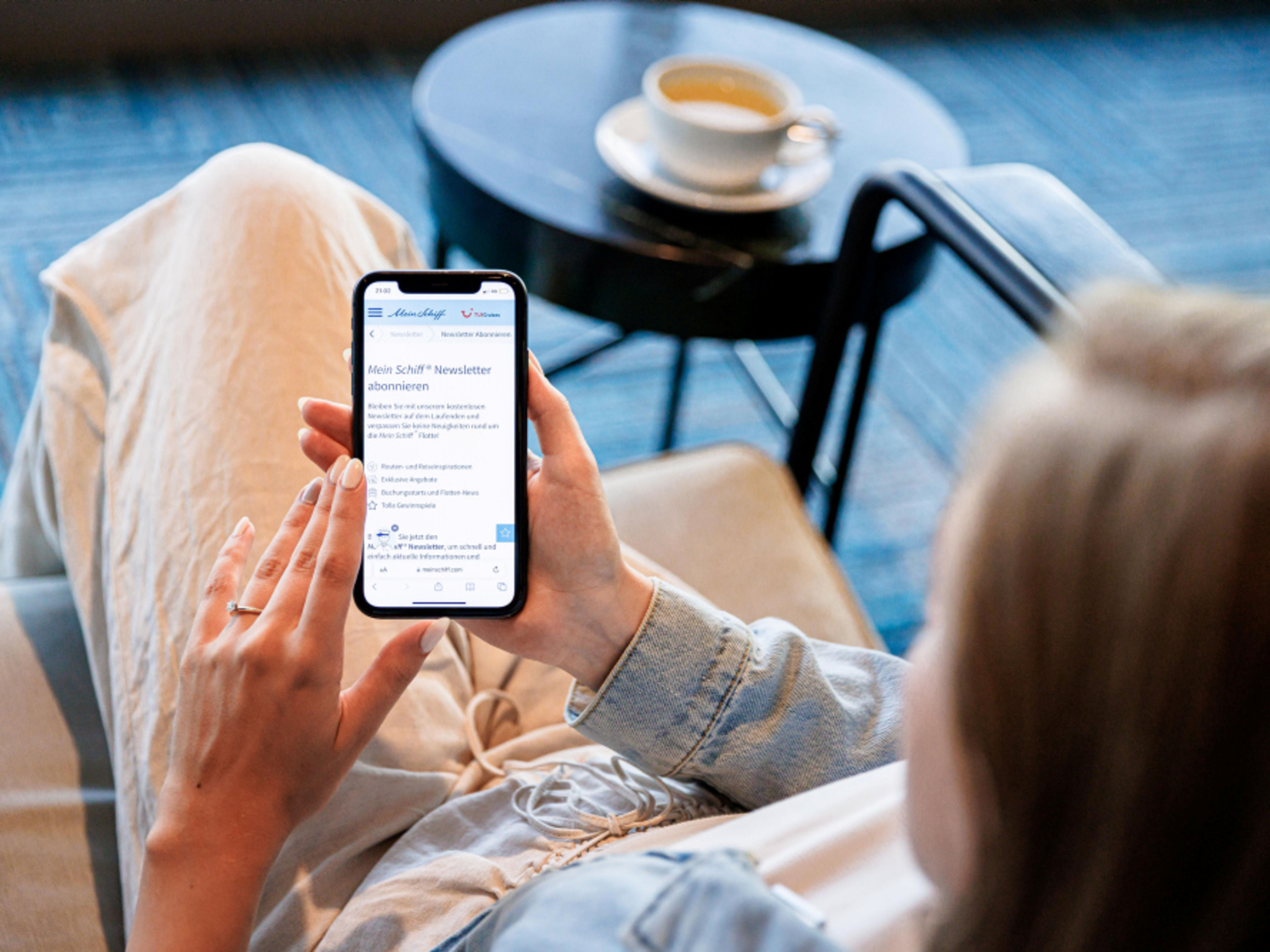Frau liest Mein Schiff® Newsletter auf Smartphone, Kaffee im Hintergrund. Frau liest Mein Schiff® Newsletter auf Smartphone, Kaffee im Hintergrund.