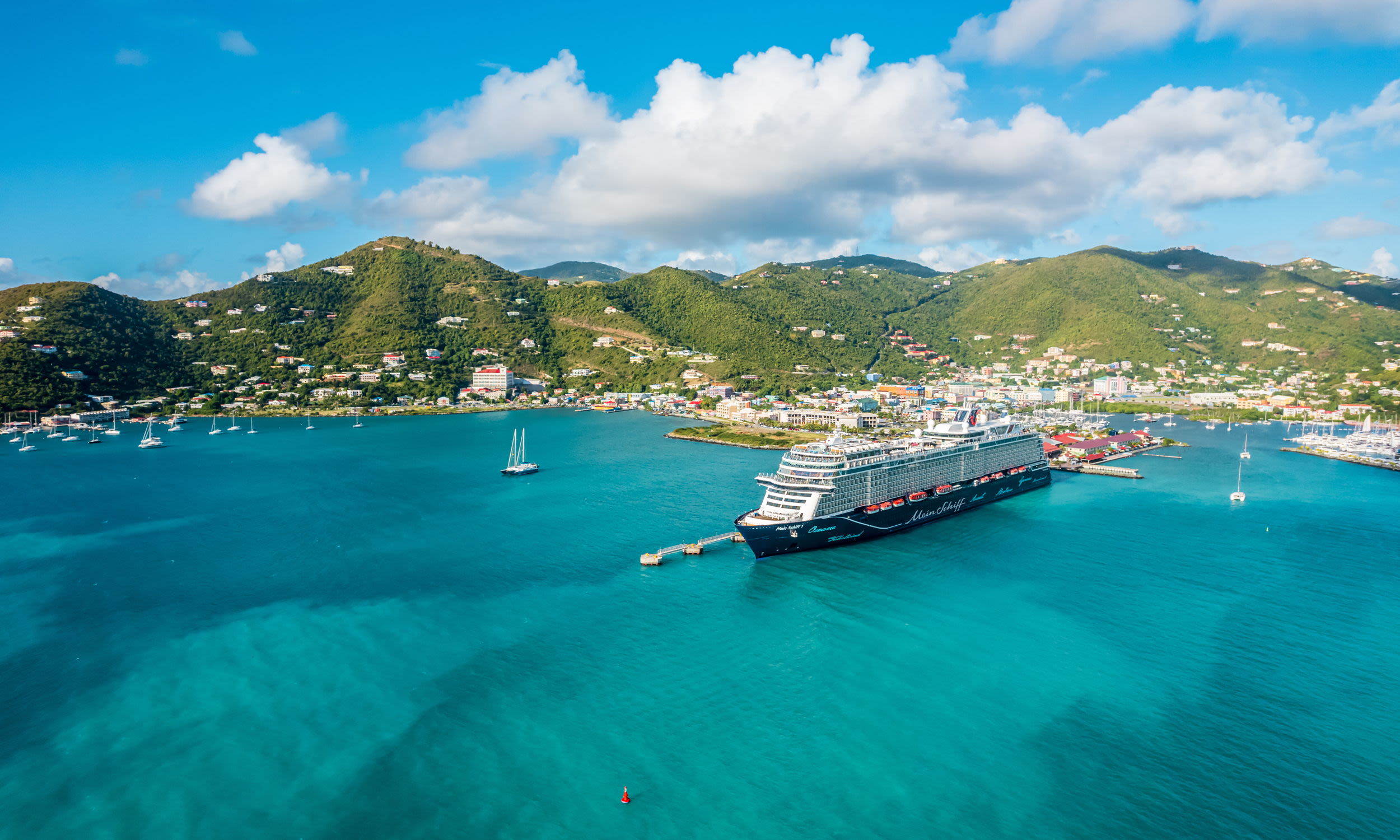Kreuzfahrtschiff der Mein Schiff® Flotte im Hafen, türkisblaues Wasser, grüne Hügel im Hintergrund.