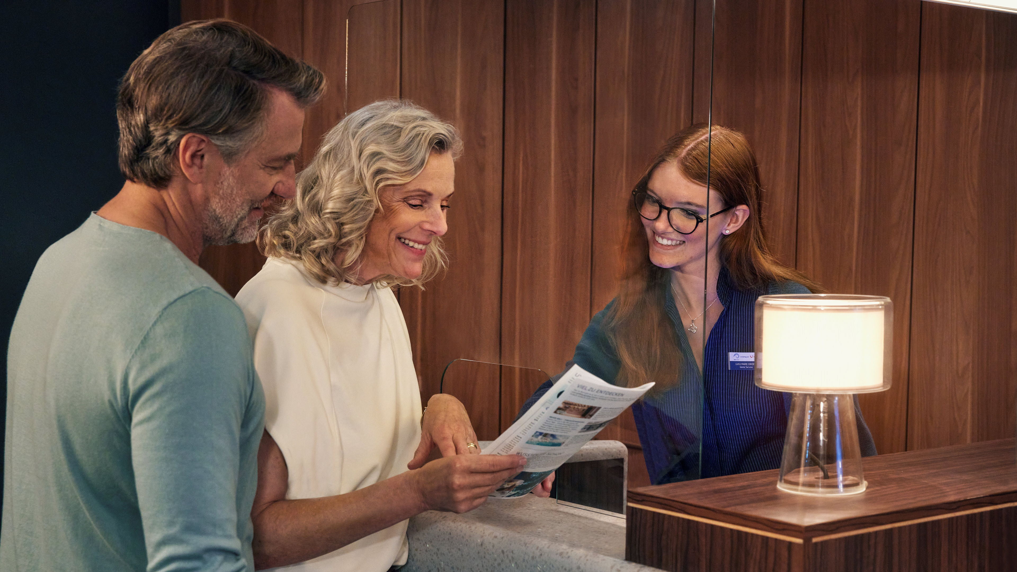 A couple at the reception desk, a smiling member of staff, wooden panelling, a lamp.