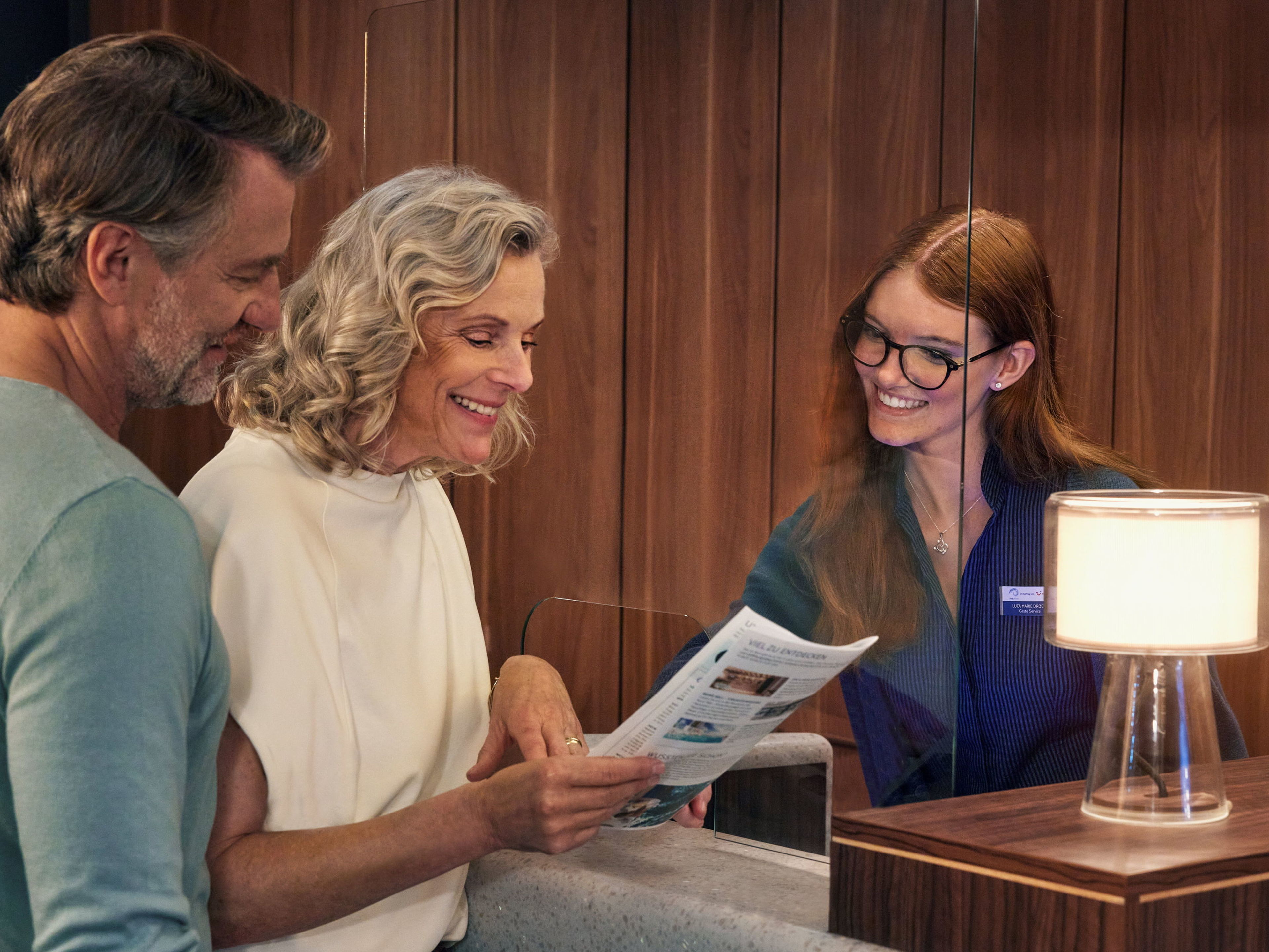 A couple at the reception desk, a smiling member of staff, wooden panelling, a lamp. A couple at the reception desk, a smiling member of staff, wooden panelling, a lamp.