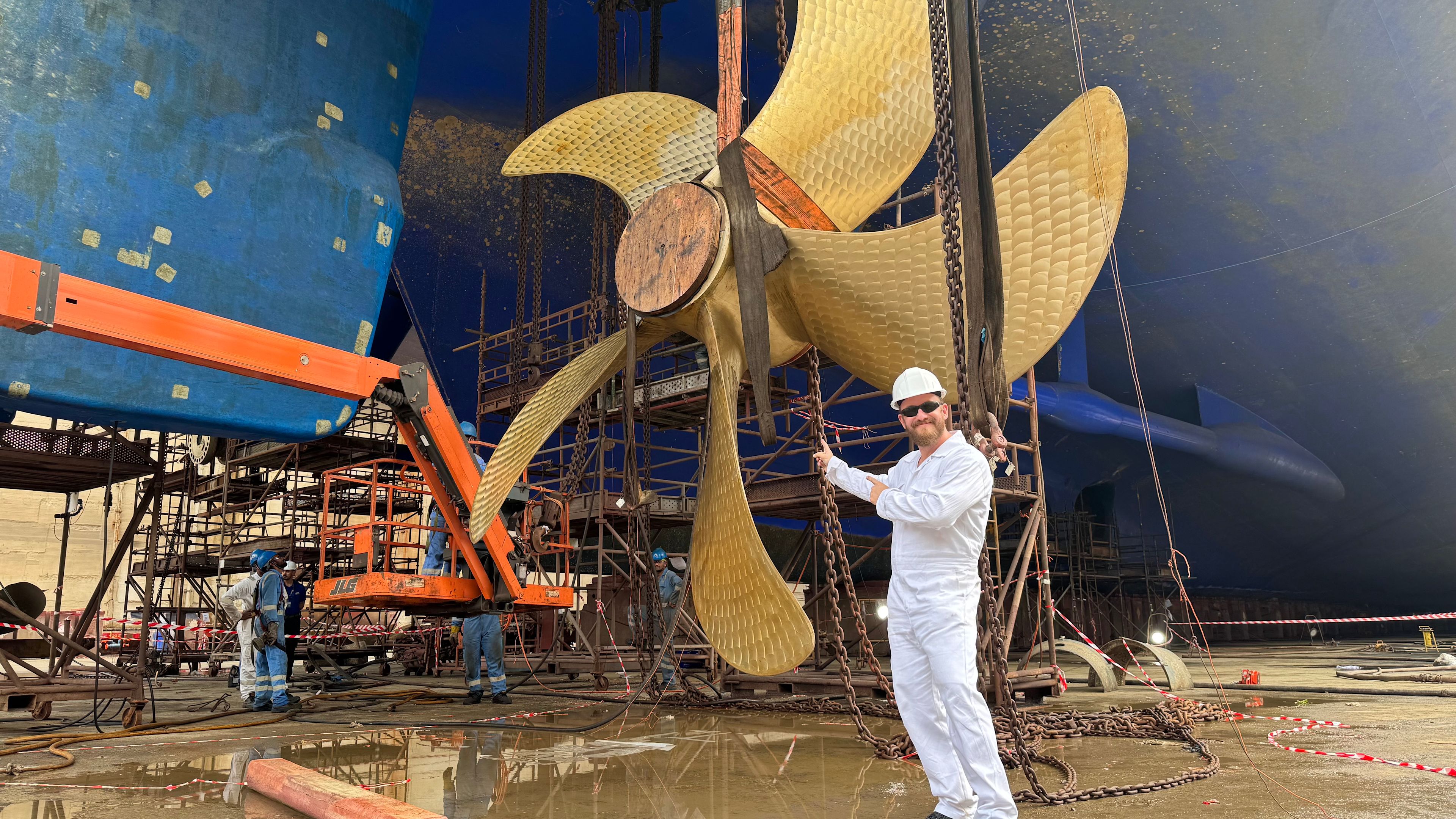 Mann in weißem Overall vor großem Schiffspropeller in Werft. Mann in weißem Overall vor großem Schiffspropeller in Werft.