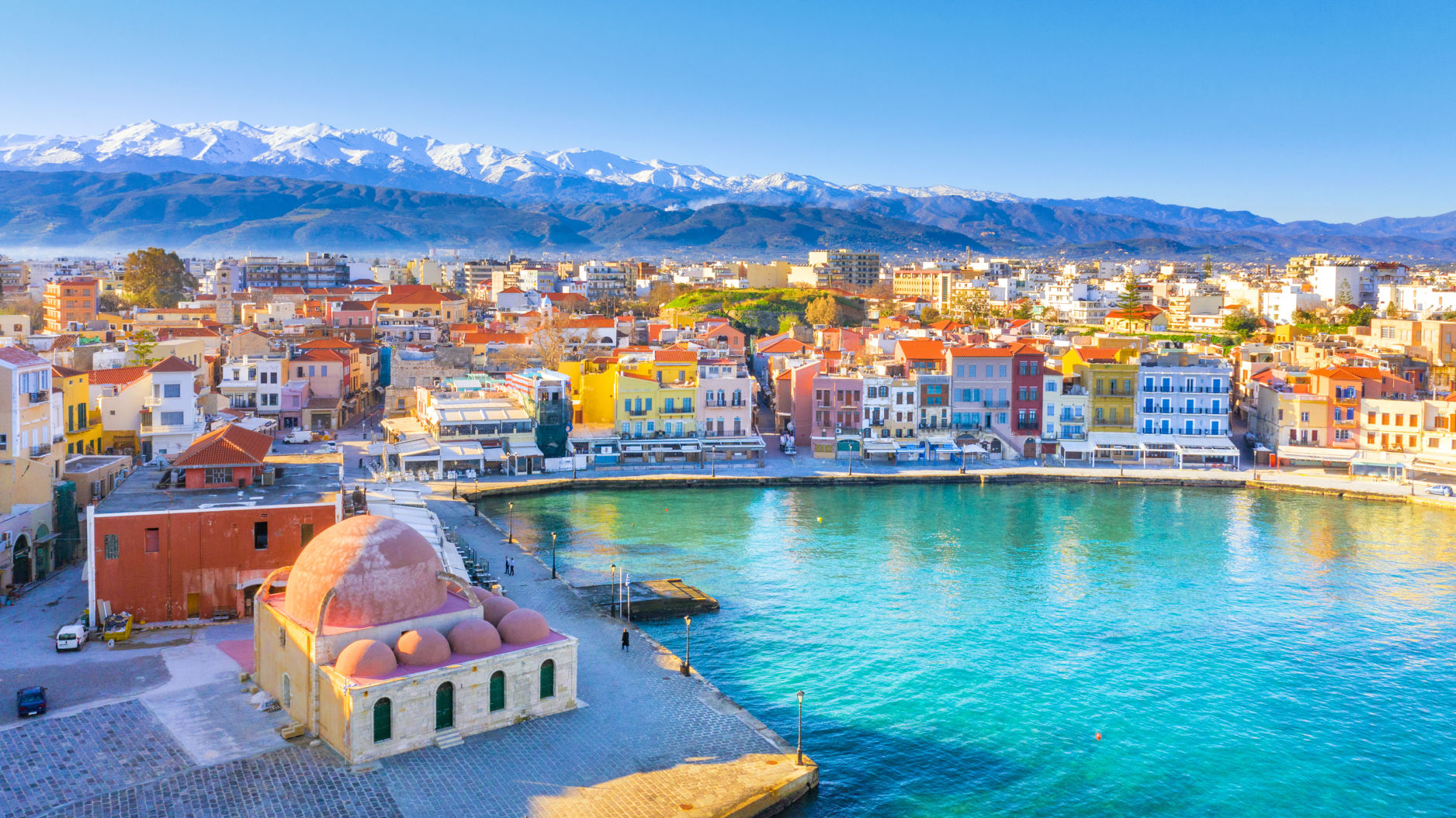 Bunte Häuser am Hafen von Chania, Kreta, mit schneebedeckten Bergen im Hintergrund.