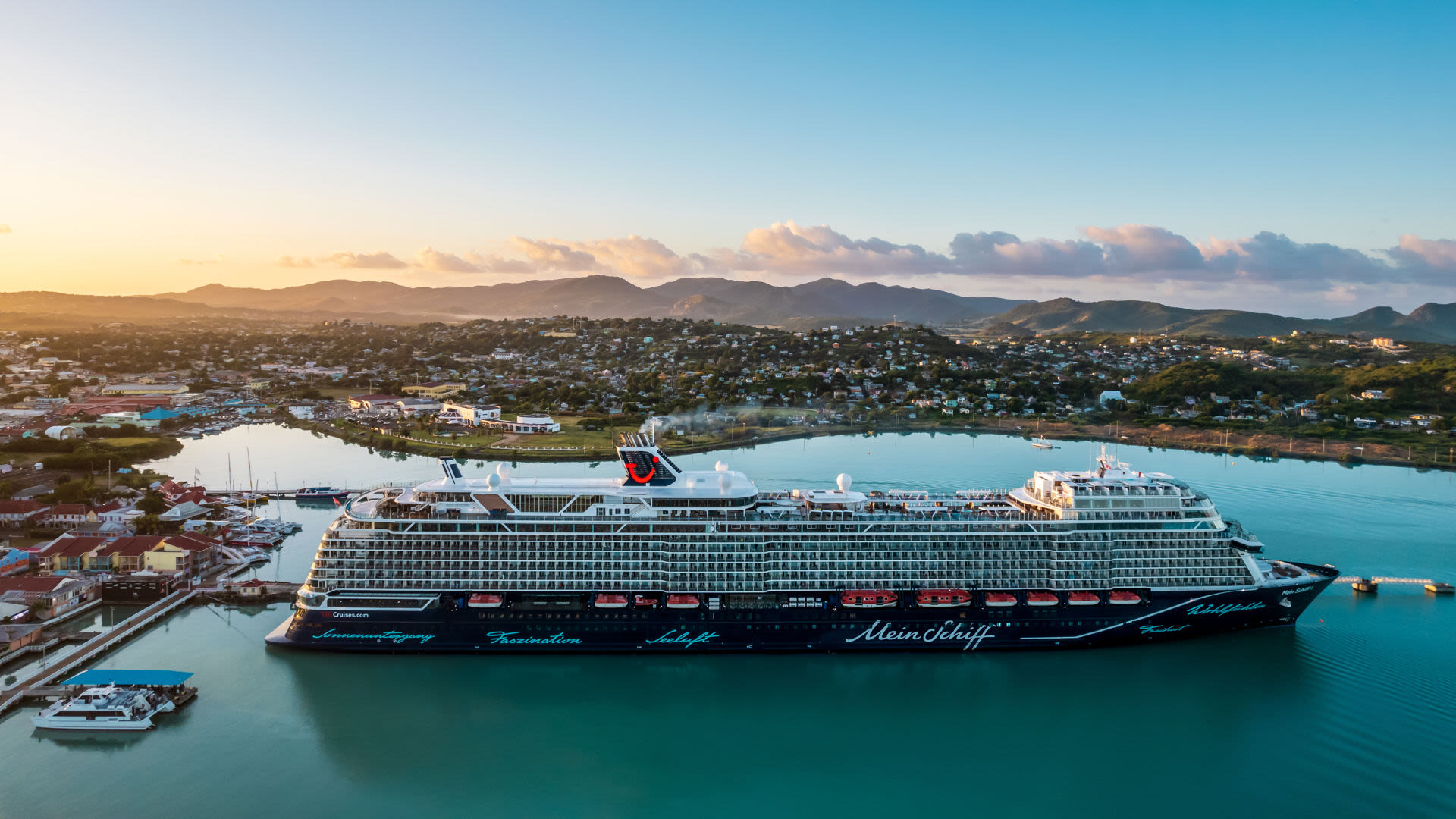 Kreuzfahrtschiff der Mein Schiff® Flotte im Hafen bei Sonnenuntergang, umgeben von Bergen und Stadt.