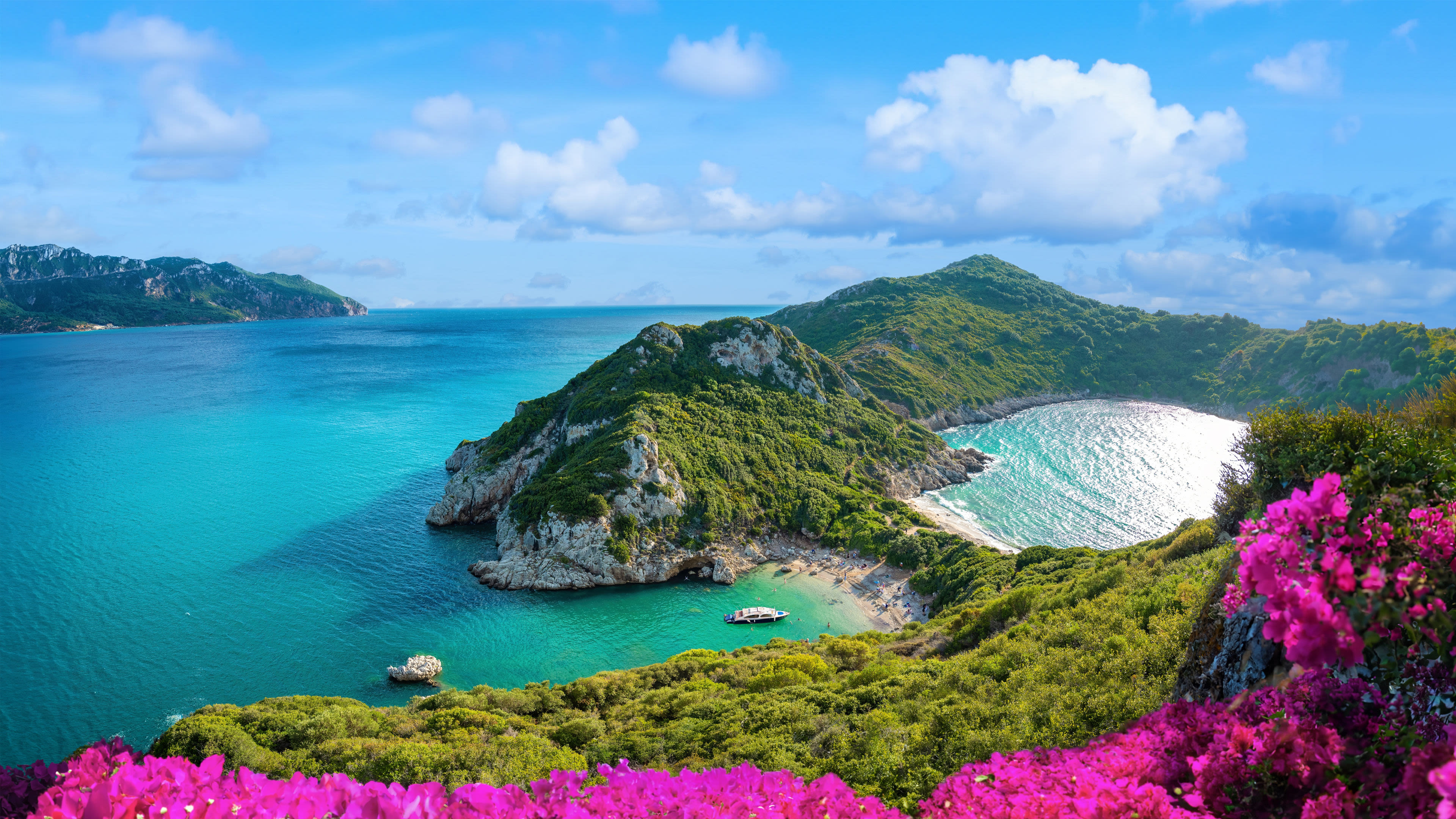 Küste von Korfu mit türkisfarbenem Meer, grünen Hügeln und pinken Blumen.