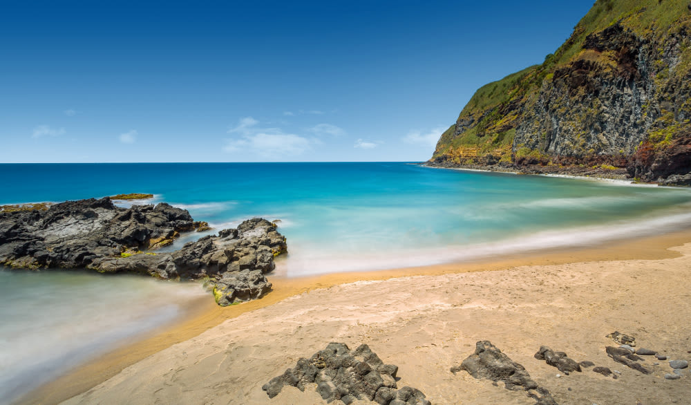 Sandstrand mit Felsen, blauem Meer und grüner Klippe unter klarem Himmel. Sandstrand mit Felsen, blauem Meer und grüner Klippe unter klarem Himmel.