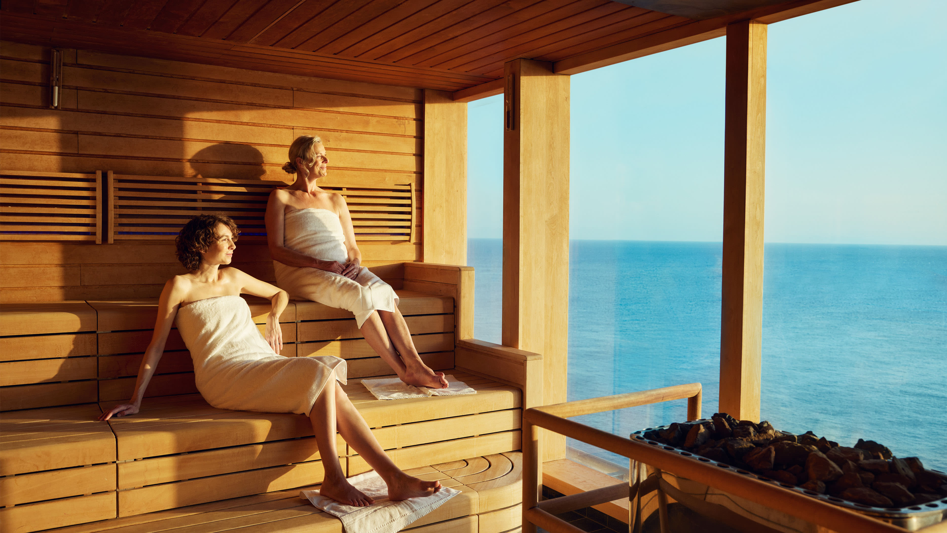 Two women relax in a wooden sauna, with sweeping sea views through large floor-to-ceiling windows. Two women relax in a wooden sauna, with sweeping sea views through large floor-to-ceiling windows.