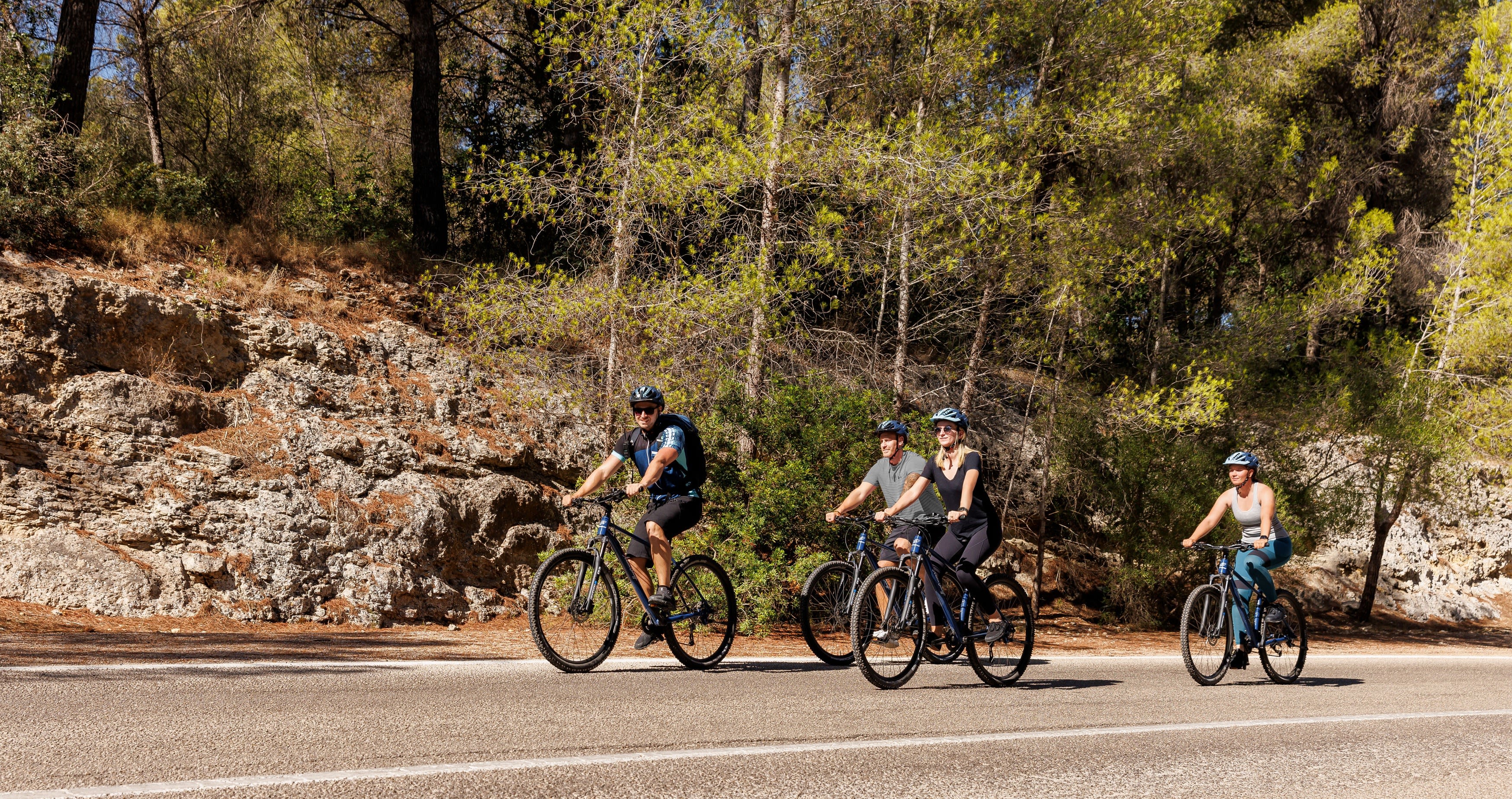 Vier Radfahrer auf einer Straße im Wald, sonniger Tag.
