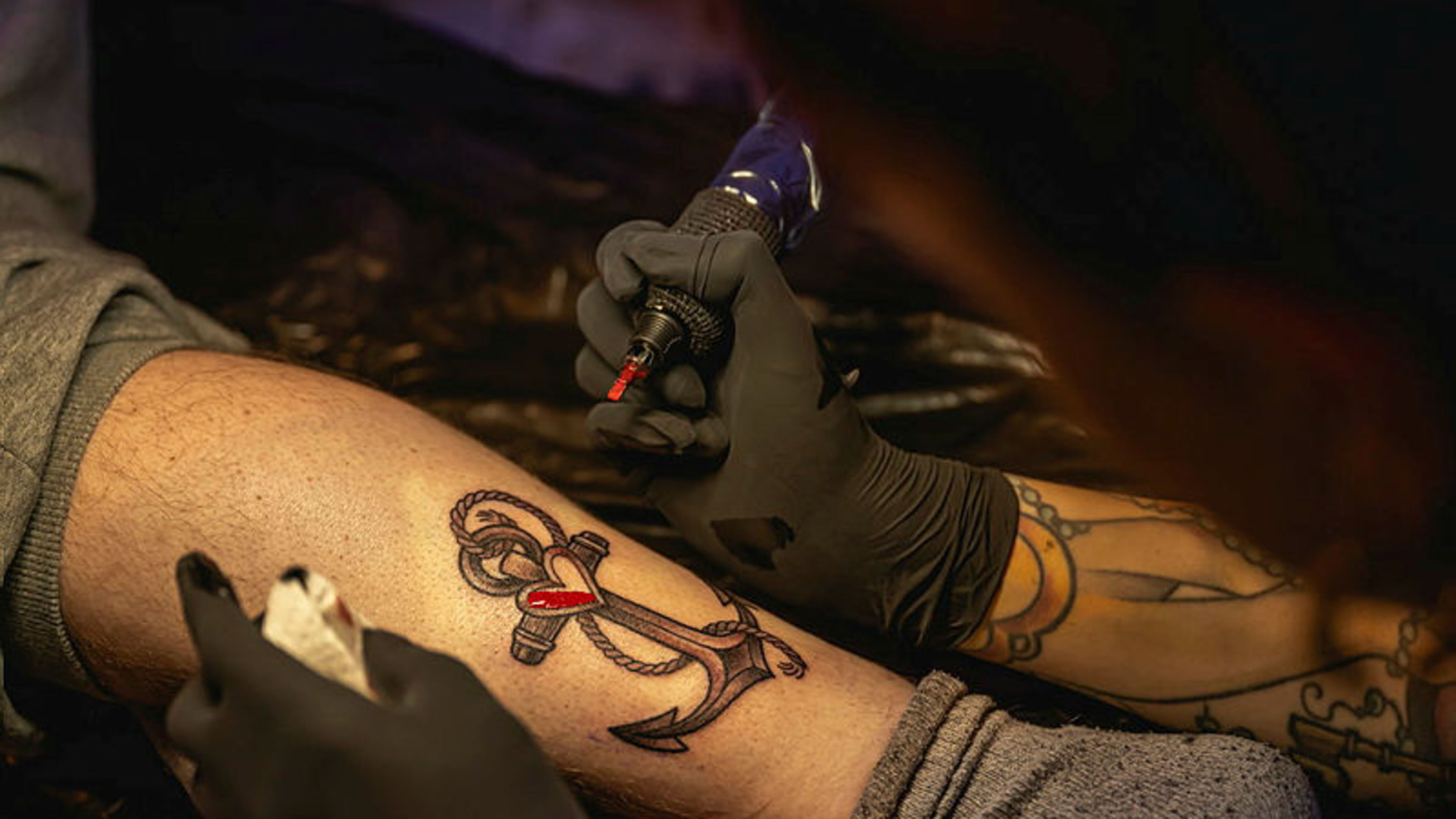 Tätowierer sticht Anker-Tattoo mit Herz auf Bein. Tätowierer sticht Anker-Tattoo mit Herz auf Bein.