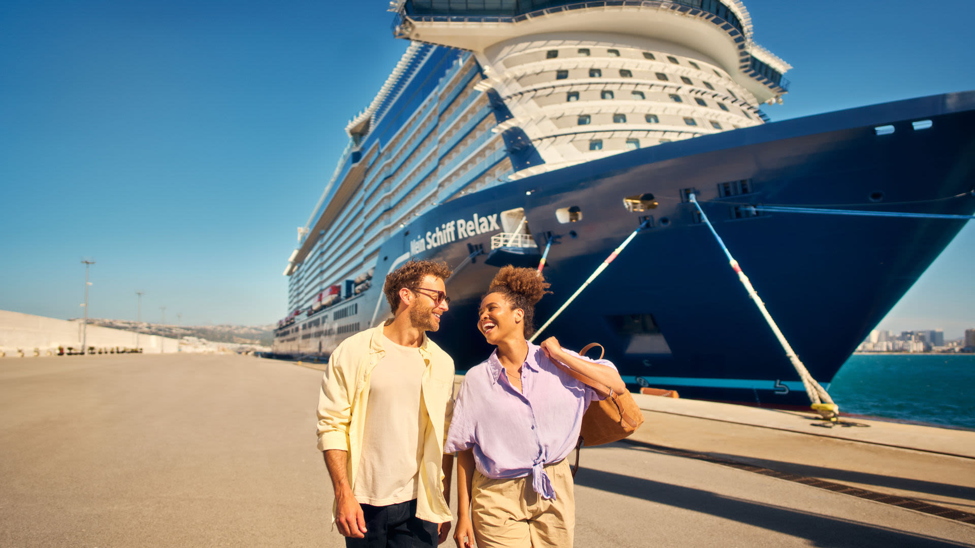 Paar spaziert vor Kreuzfahrtschiff der Mein Schiff® Flotte, sonniger Tag.