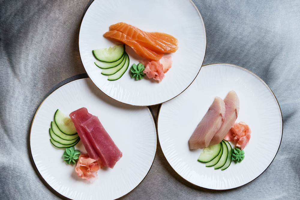 Drei Teller mit Sashimi, Gurken, Wasabi und Ingwer auf grauem Hintergrund.